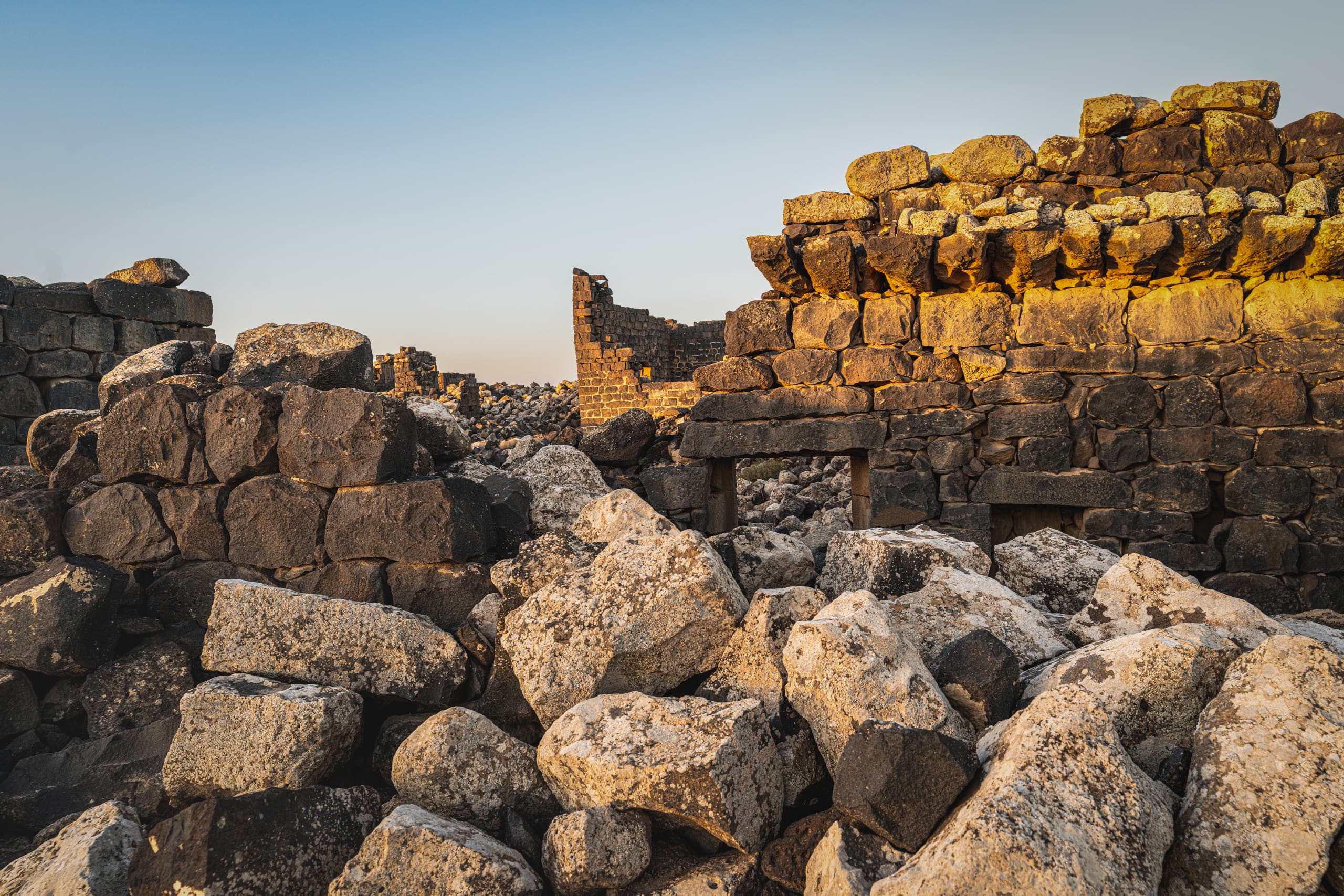 A low-angle view of crumbling basalt stone walls and a pile of large, weathered boulders under a clear sky, illuminated by golden hour light.