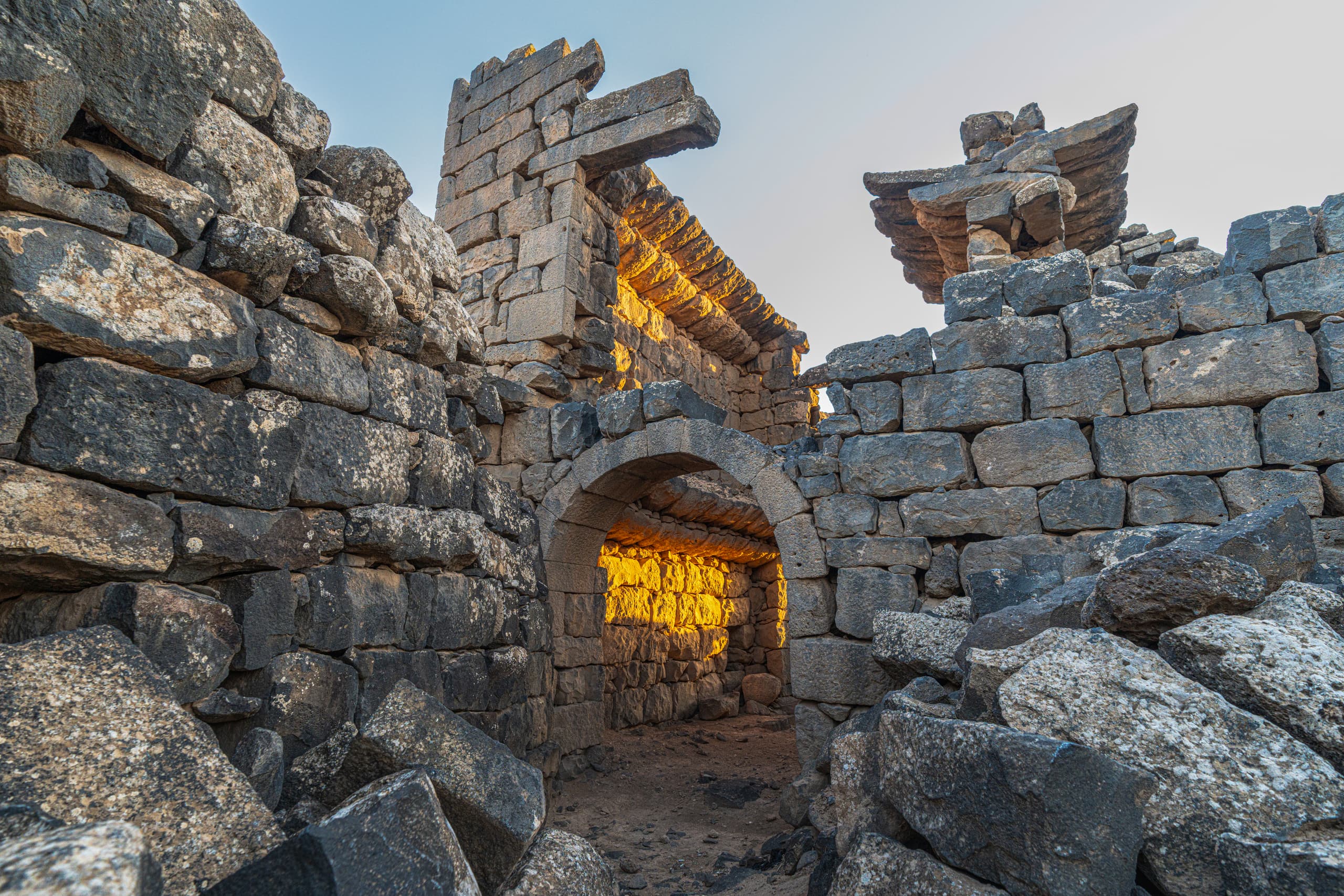 Low-angle shot of ancient stone ruins and a rounded archway illuminated by golden sunset light against a pale sky.
