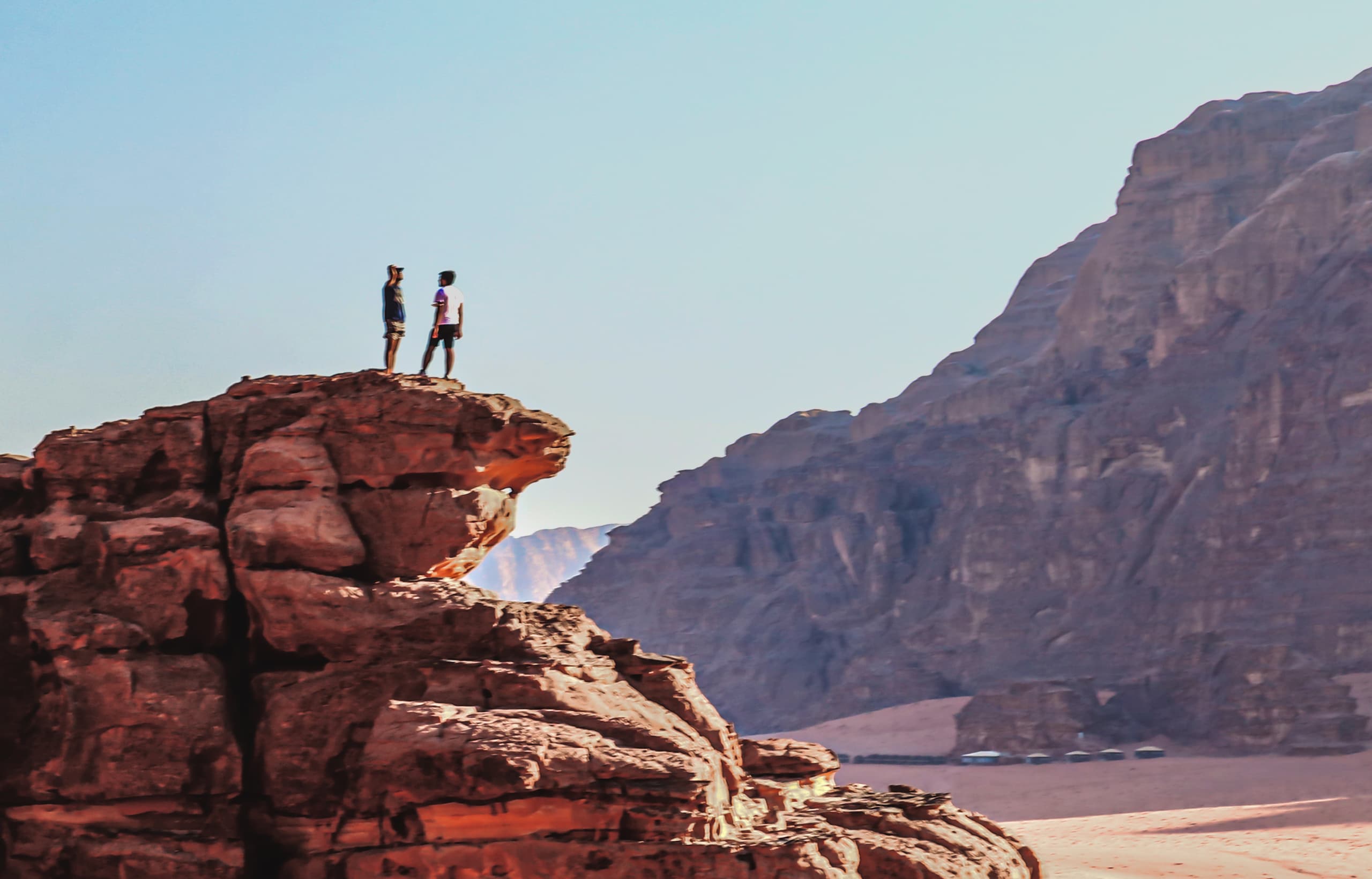 Two people standing on the rocky outcrop of a desert cliff, viewed against a vast expanse of mountains and clear sky.