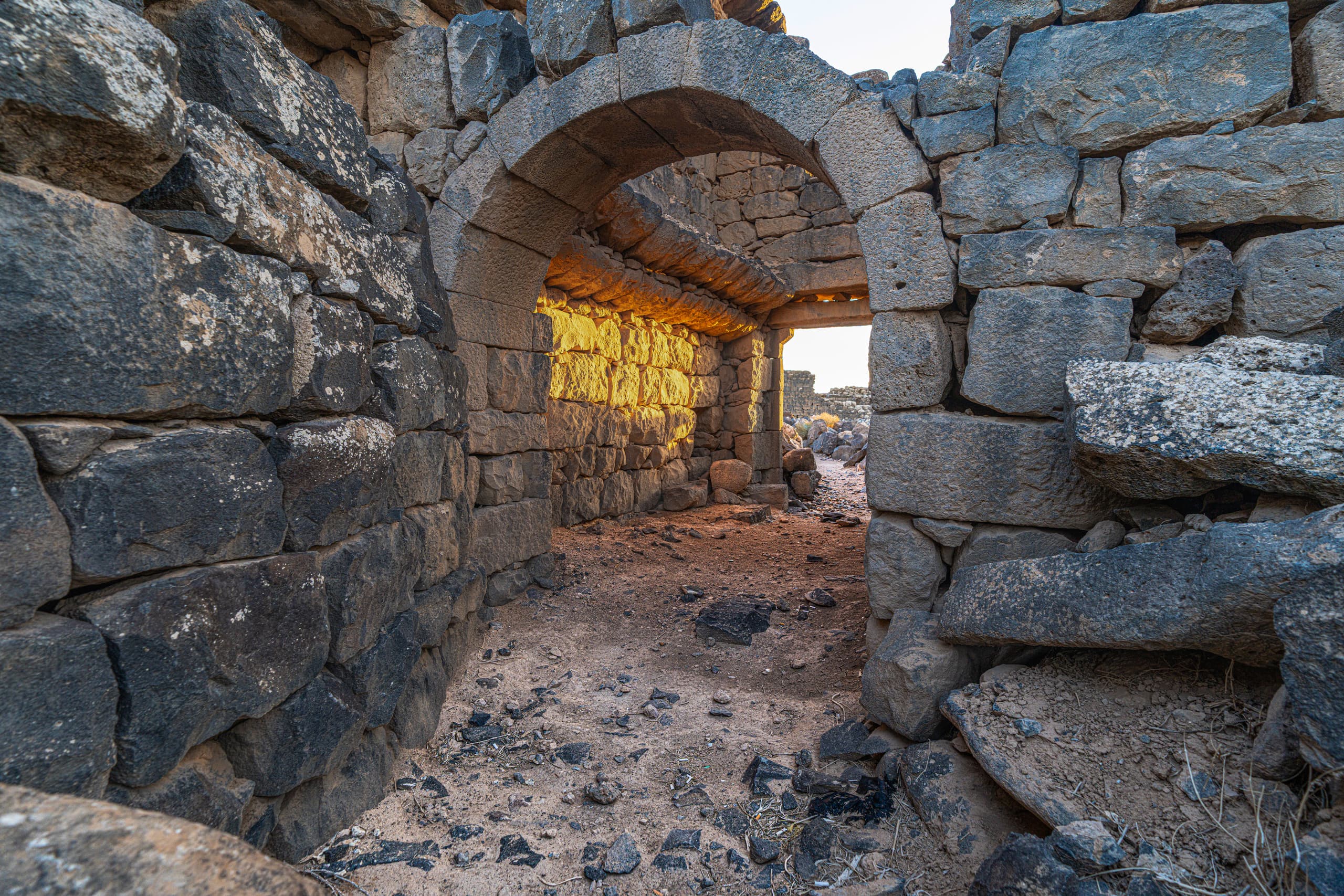 Interior view through a stacked basalt stone archway and passageway, highlighting rough rock textures and warm golden evening light hitting the back interior wall.