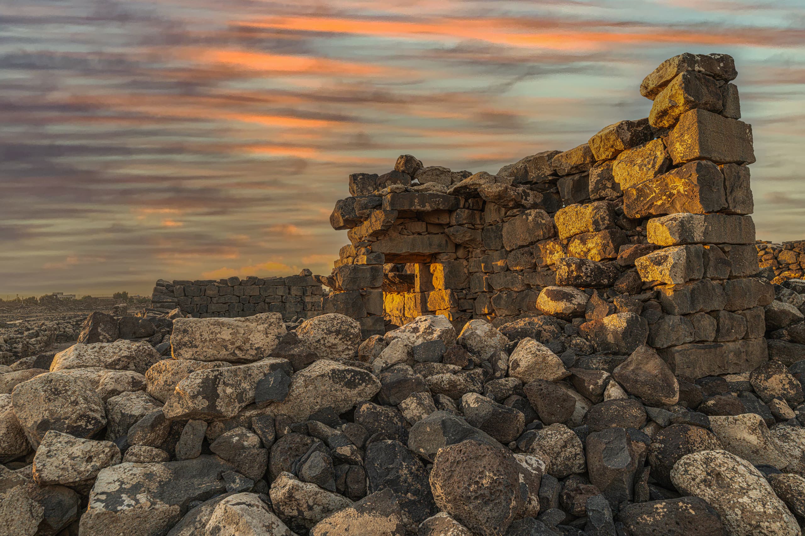 Ancient stone ruins composed of stacked dark basalt blocks and scattered boulders in the foreground, illuminated by golden hour light under a striated sunset sky.
