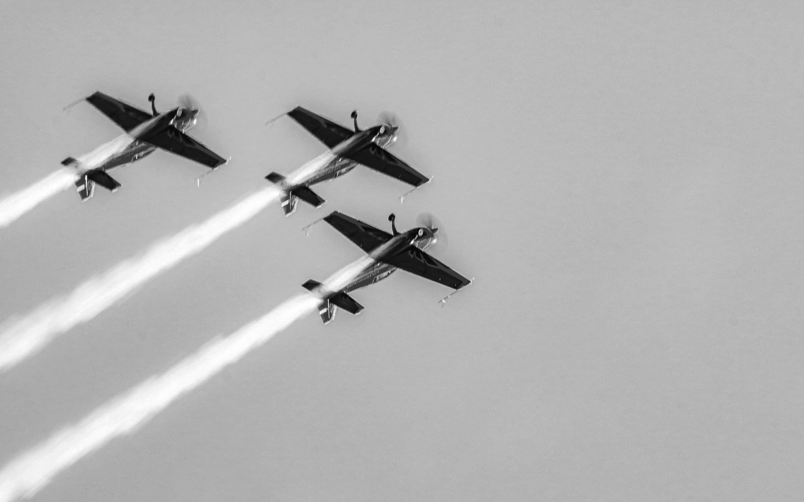 A black-and-white low-angle shot showing three small propeller planes flying in diagonal formation from upper left toward lower right, leaving heavy white vapor trails across a uniform sky.