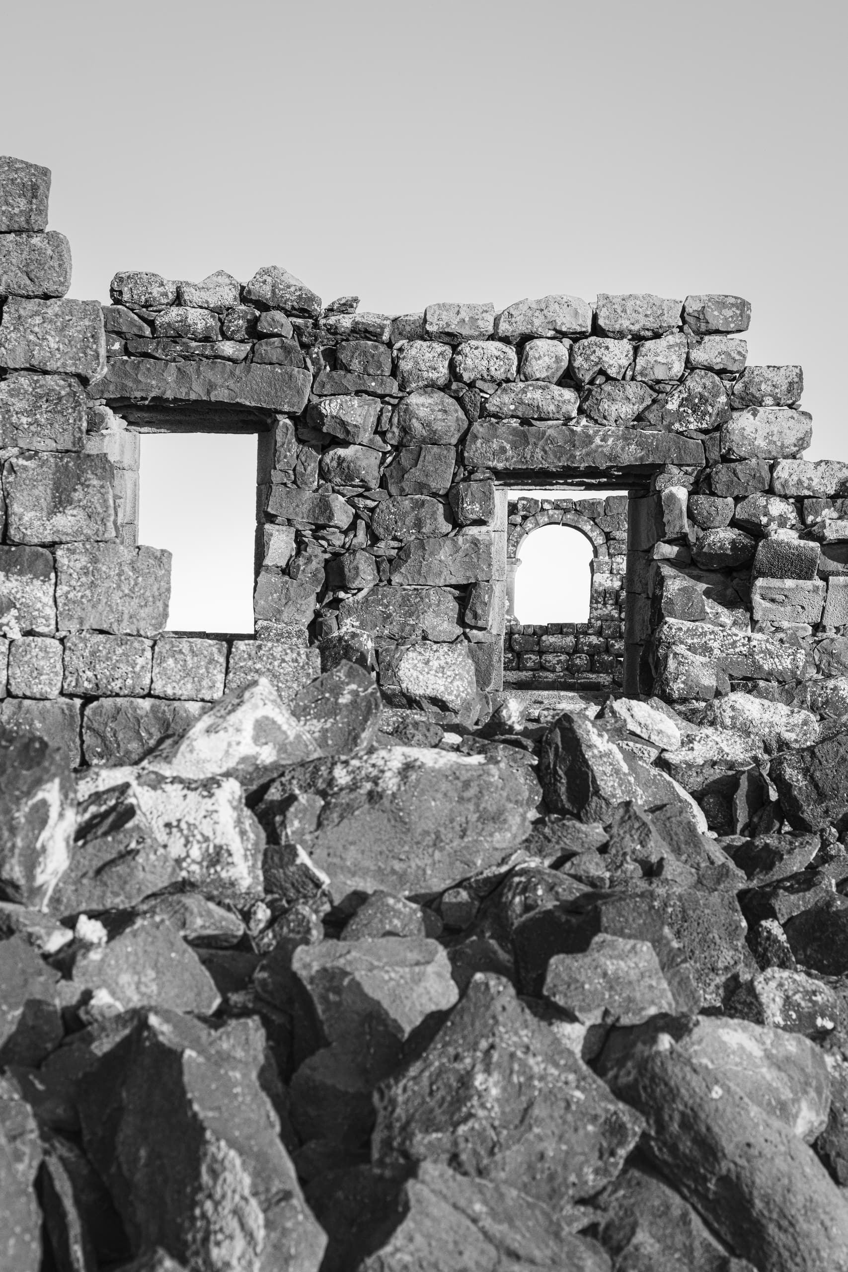 Monochrome architectural photograph showing a ruined stone wall made of dark basalt blocks, featuring two square window openings and an arched doorway framing a bright sky in the background.
