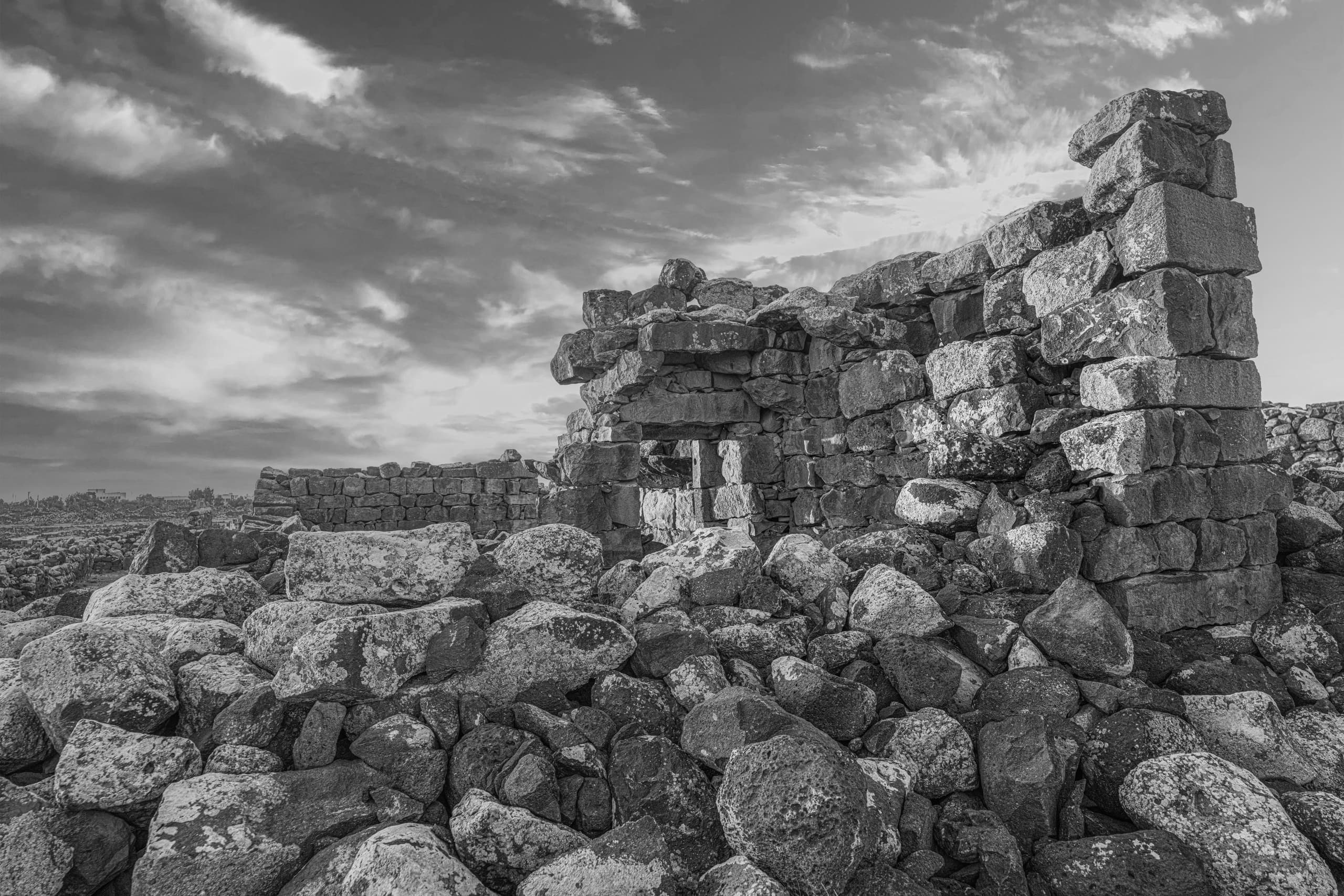 Low-angle monochrome photograph of weathered stone ruins and piled basalt rocks beneath a cloudy sky.
