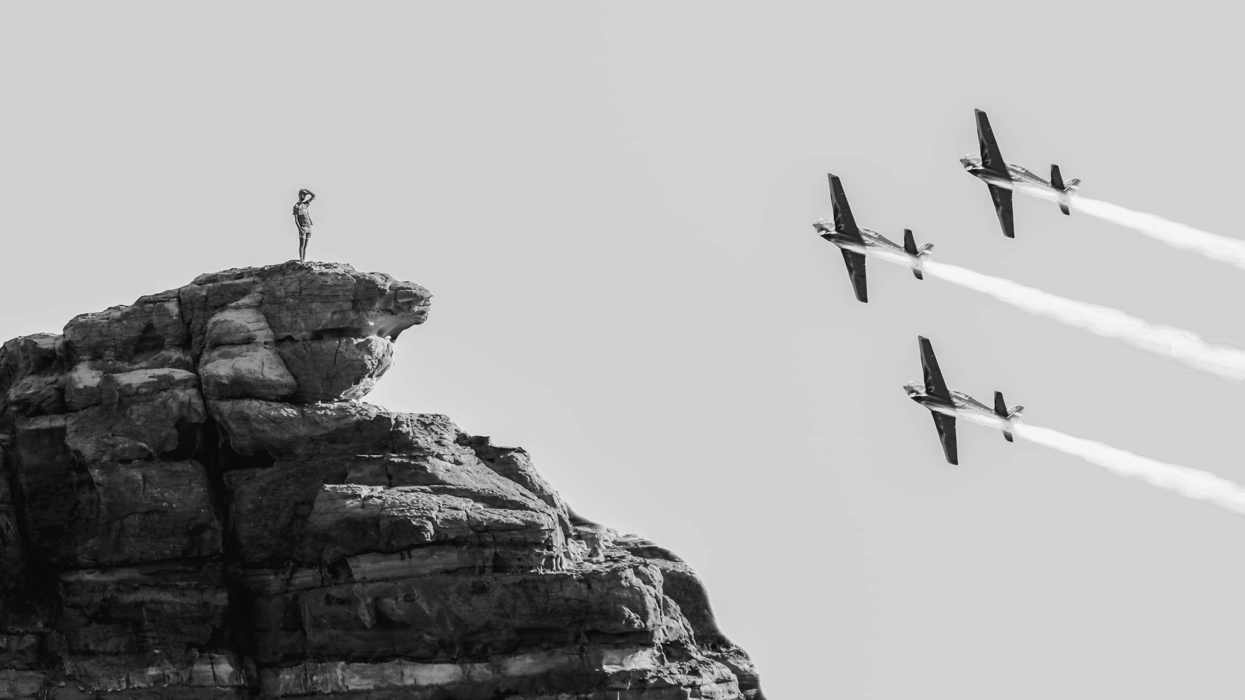 Monochrome image of a person standing on top of a rocky cliff looking at three airplanes flying in formation with vapor trails on the right.