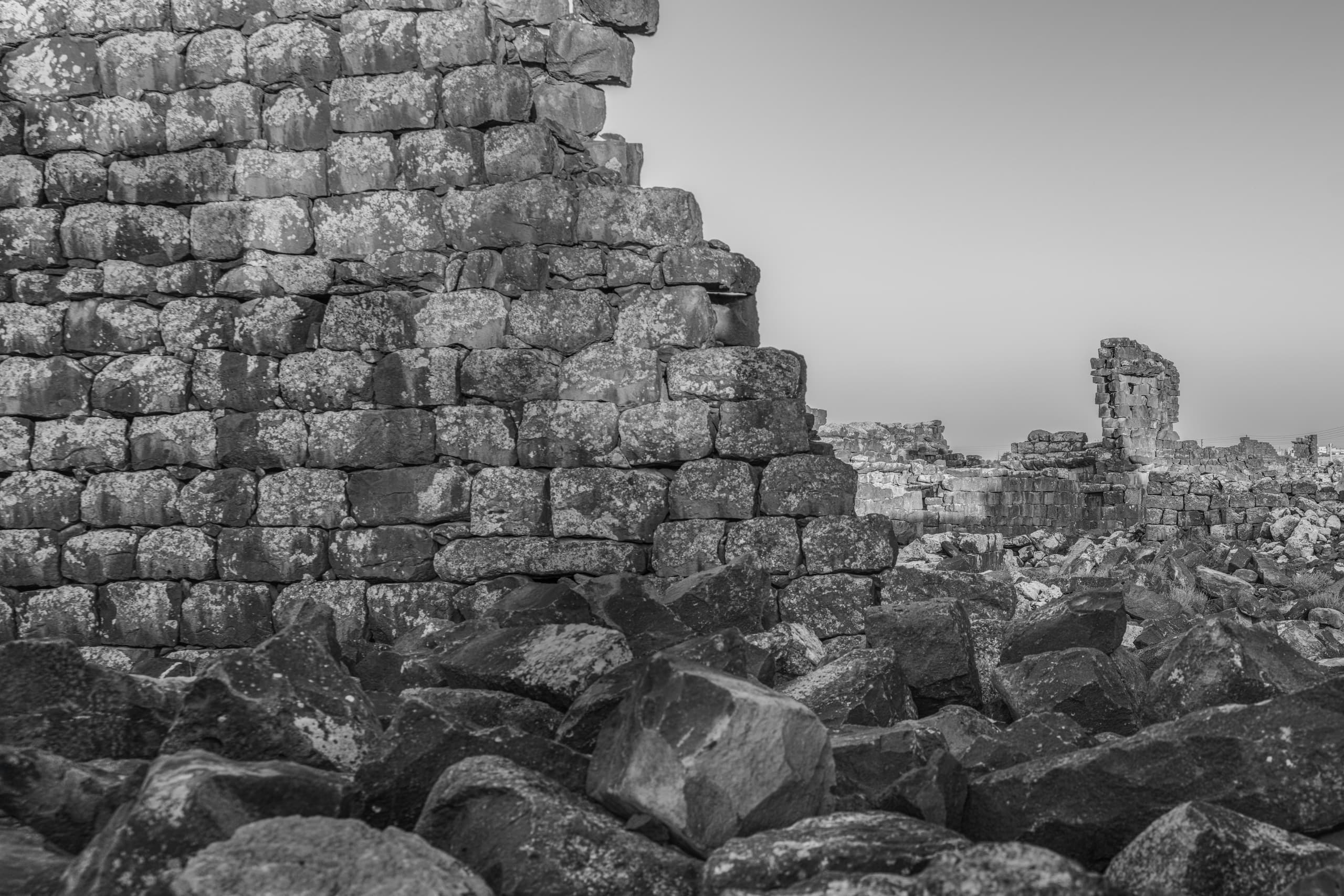 Monochrome architectural photograph showing a tall, weathered dark stone wall with lichen patches on the left, an expanse of tumbled basalt boulders in the foreground, and distant ruined structures against a clear sky.