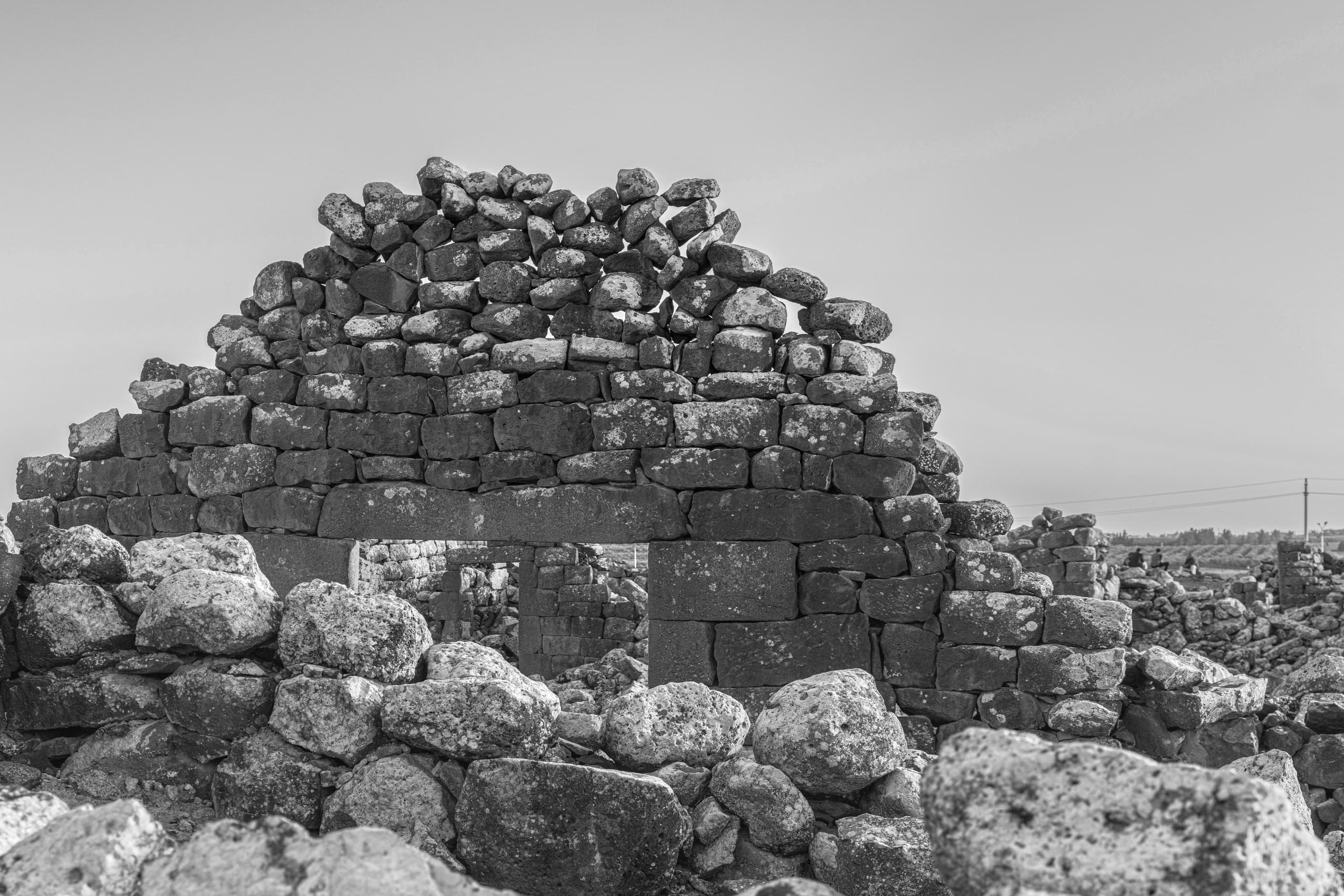 Monochrome photograph of an ancient, weathered stone structure featuring rough-hewn stacked boulders and a lintel doorway against a pale, even-toned sky.