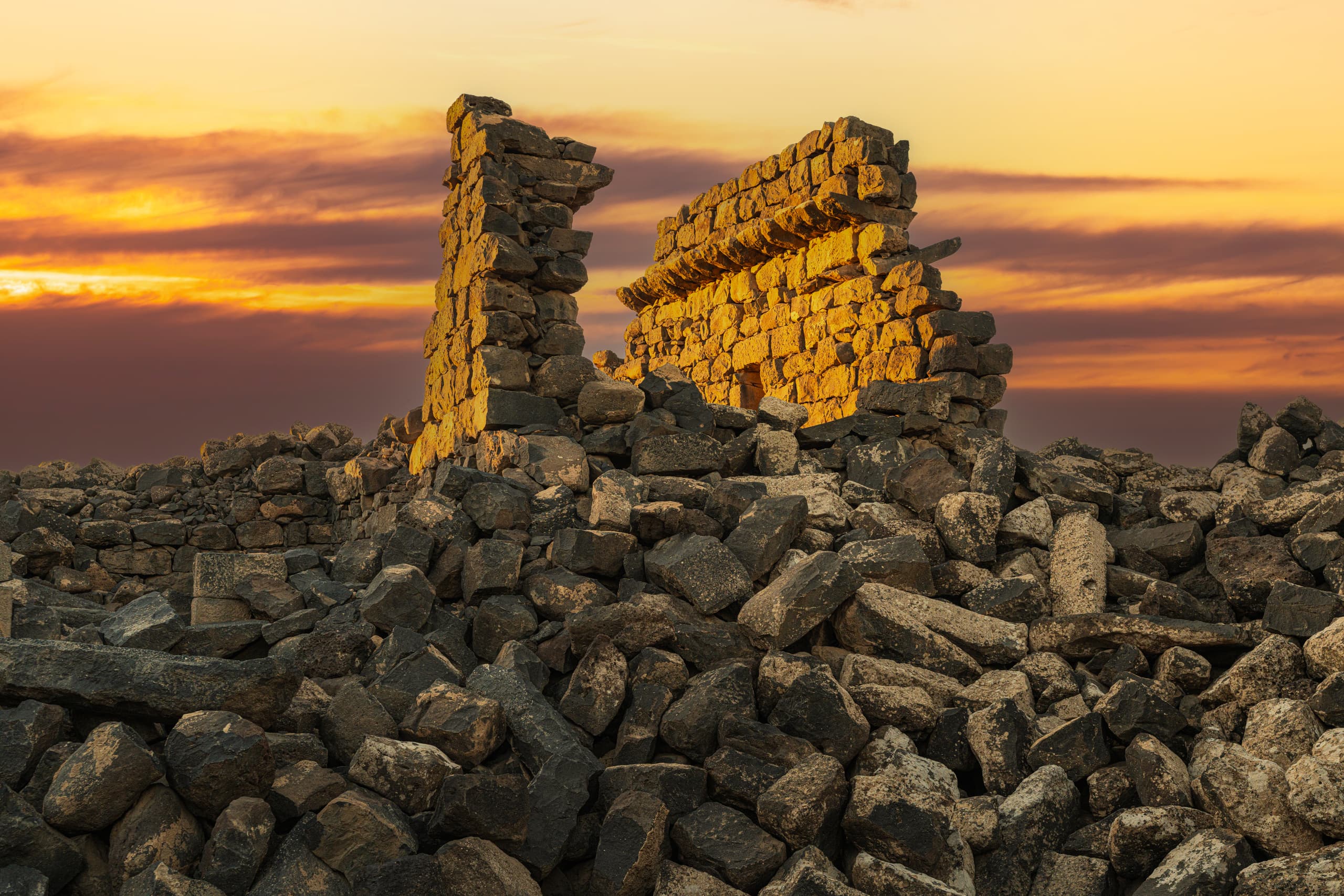 Ancient stone ruins standing atop a mound of dark rubble against a colorful golden sunset sky with layered clouds.