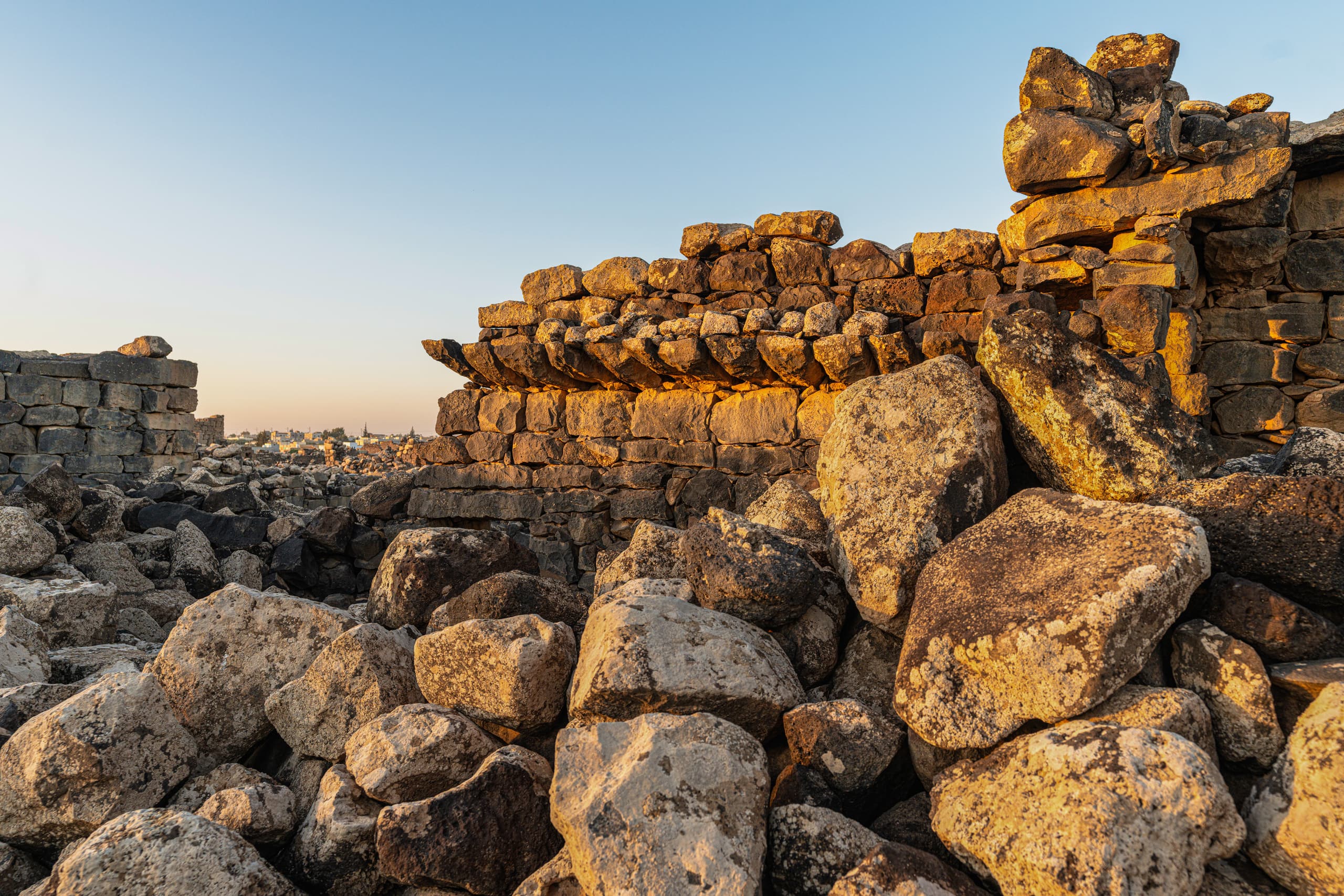 A low-angle view of rugged basalt boulders piled in the foreground, with an ancient stacked-stone ruin illuminated by golden late-afternoon sunlight under a clear blue sky.