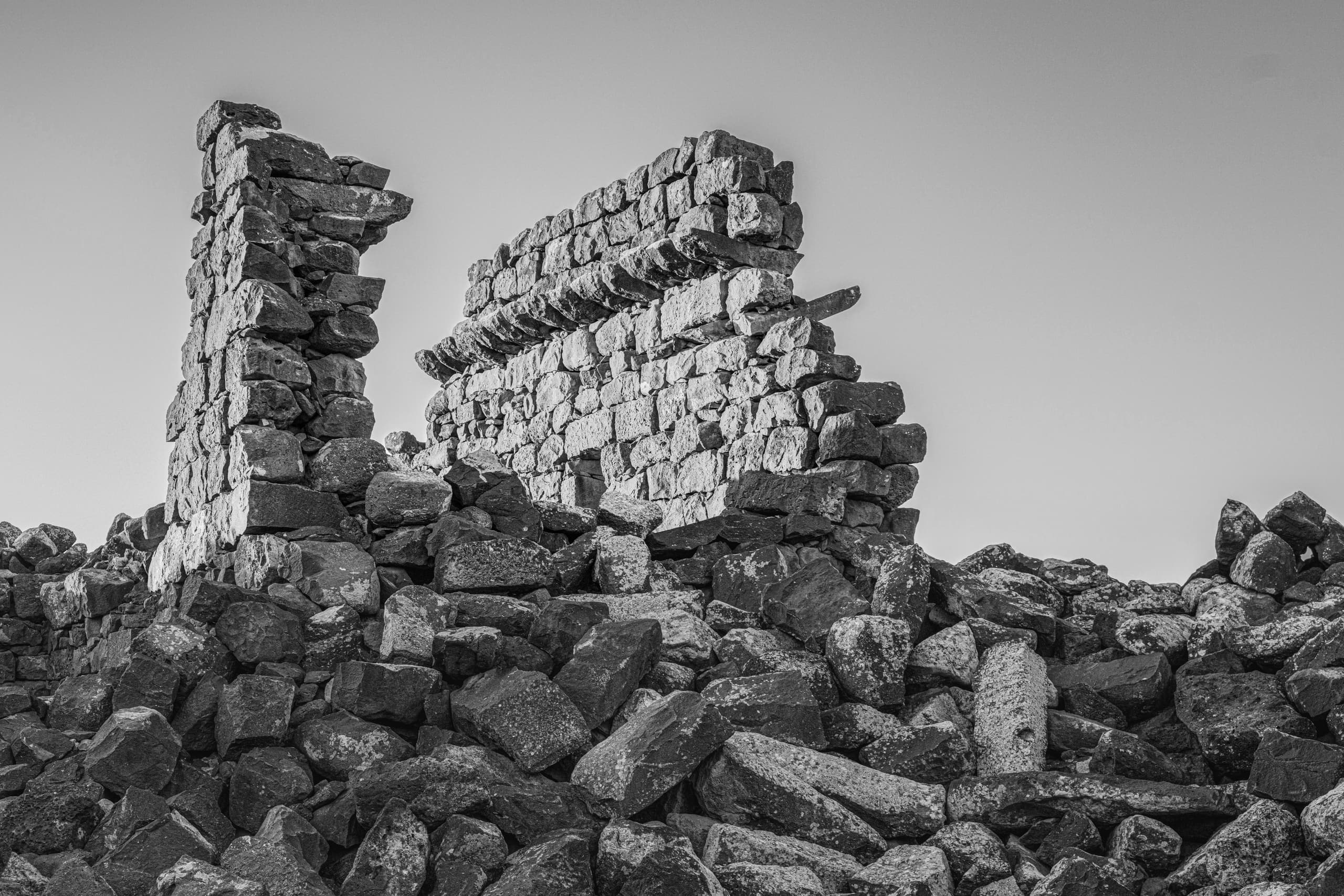 Black and white photograph of ancient stone ruins and rubble with dark basalt blocks piled in the foreground against a pale sky.