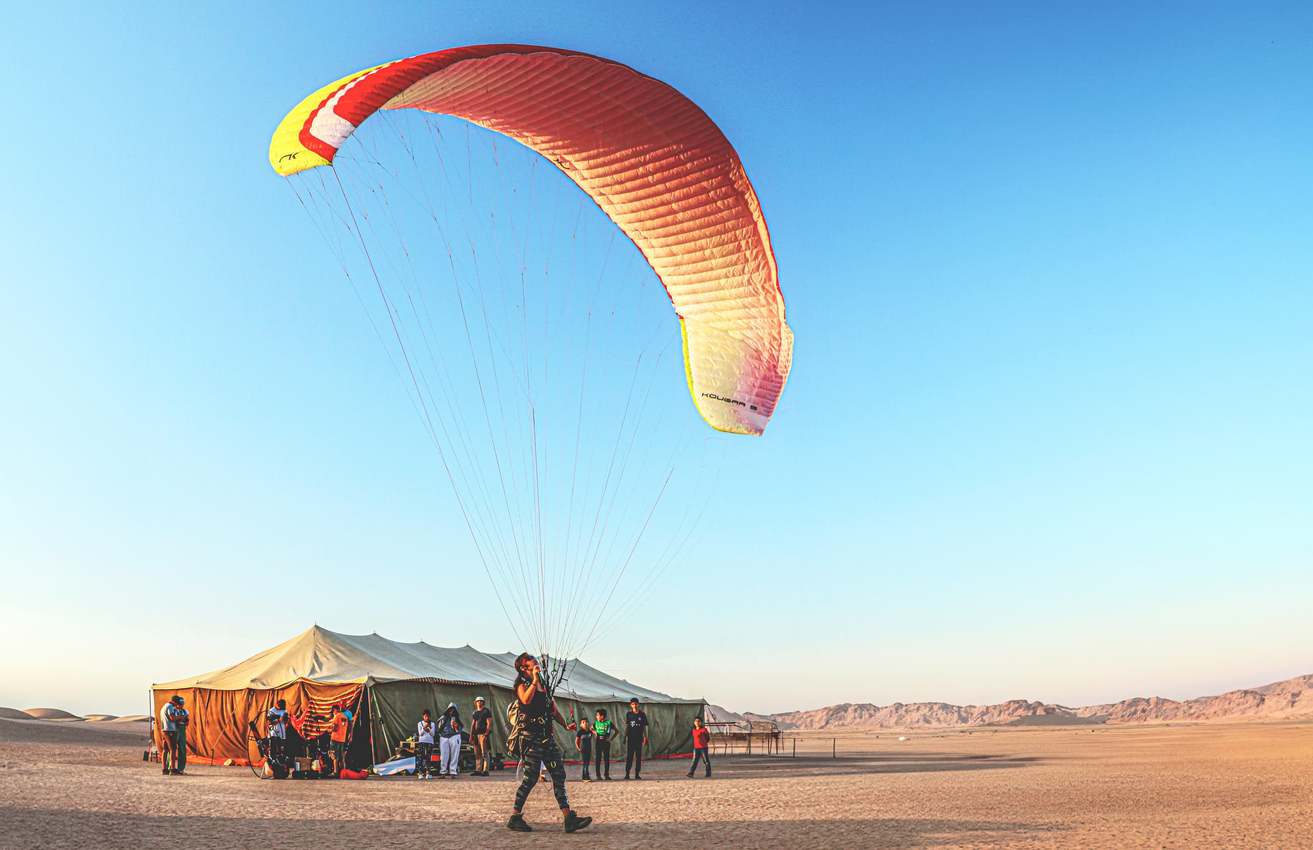 A paraglider in dark gear stands holding the lines of a red, white, and yellow paraglider wing that is inflated overhead in a vast desert landscape next to a large tent with several people standing nearby.