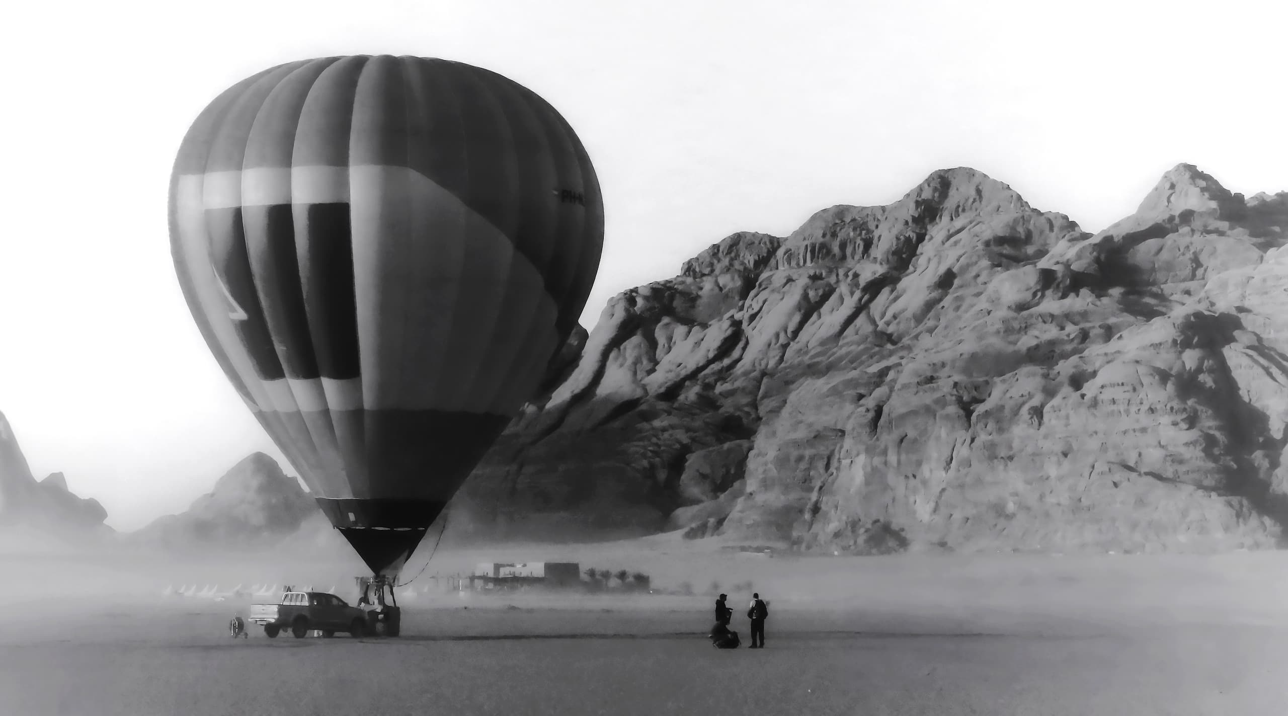 Monochrome photograph of a large hot air balloon grounded on a vast sandy plain, with two small figures standing nearby and towering rocky mountains in the background.