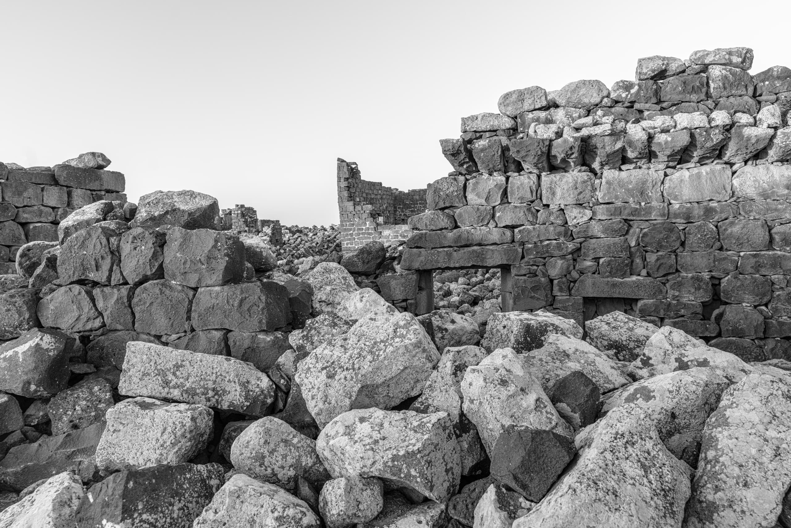 Monochrome photograph of crumbling stone ruins and jumbled basalt boulders, highlighting rough textures, broken walls, and weathered masonry against a pale sky.