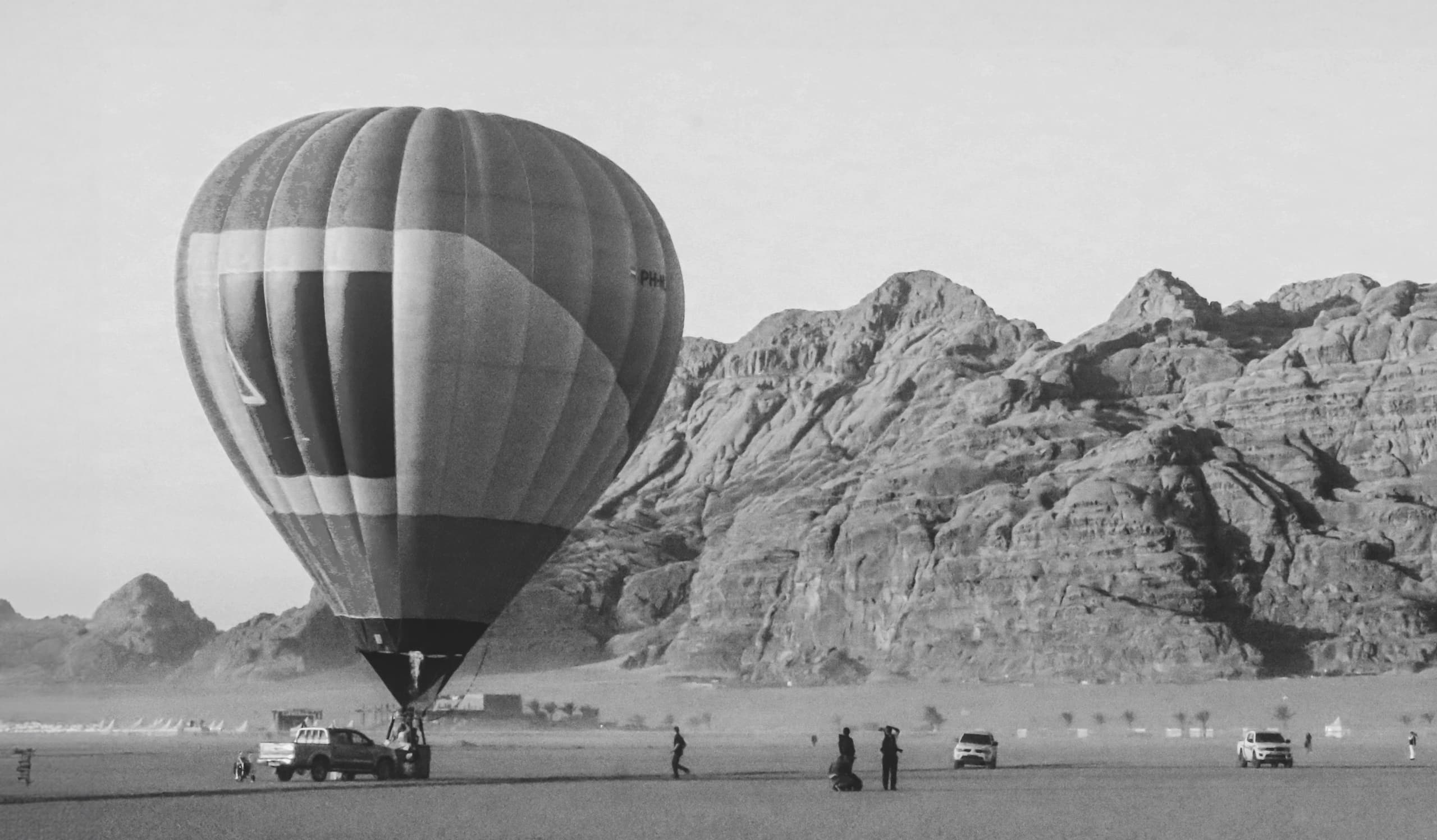 Monochrome landscape photograph featuring a large striped hot-air balloon on the left side, grounded near a pickup truck in a vast desert plain. In the background, a rugged mountain range stretches horizontally across the frame under a clear sky. Several small human figures and parked vehicles provide a sense of scale across the desert floor.