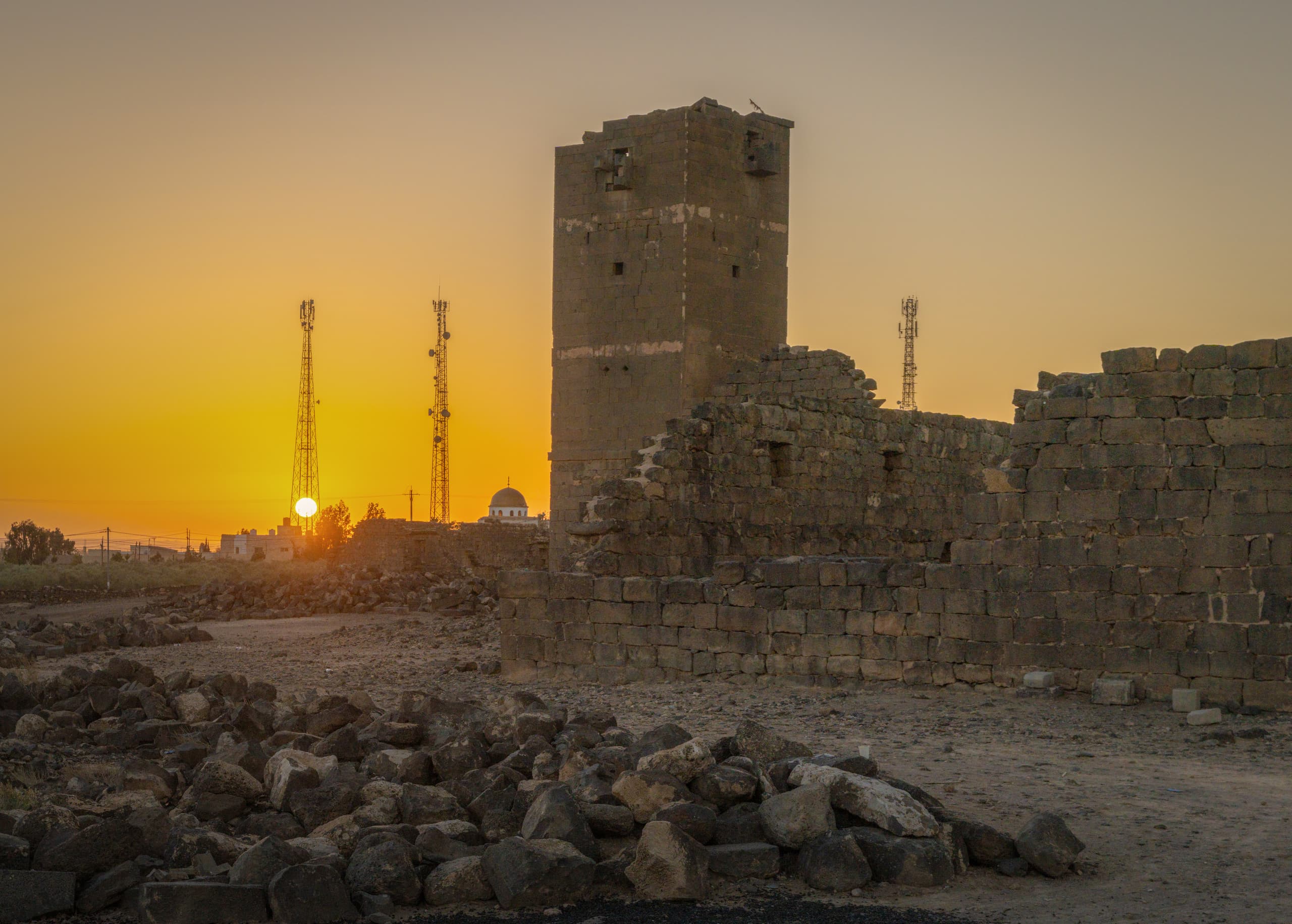 A wide-angle exterior view of ancient stone ruins and a tall weathered tower under a glowing orange sunset sky, with modern cellular towers and a distant dome visible on the horizon.