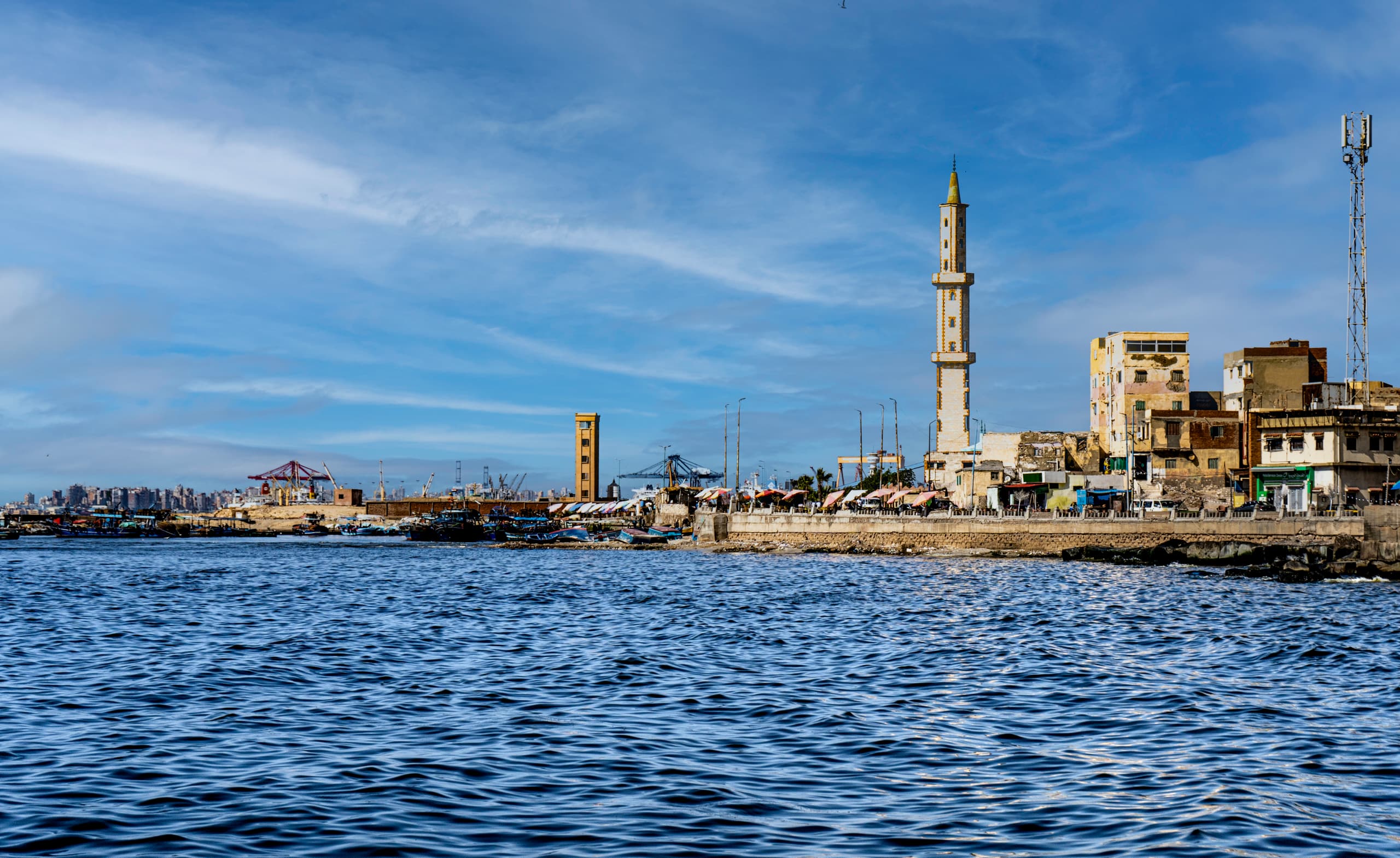 Wide exterior view of a coastal cityscape featuring a prominent tall stone minaret next to older buildings along a seawall, with choppy dark blue water in the foreground and a bright blue sky overhead.