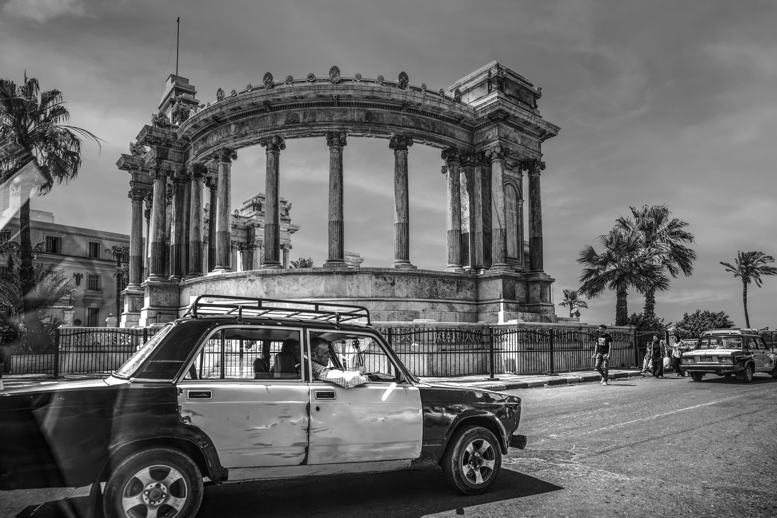Monochrome street-level photograph featuring a dark-and-light vintage taxi car moving from left to right across the foreground, with a large classical stone memorial colonnade and palm trees behind it under a cloudy sky.