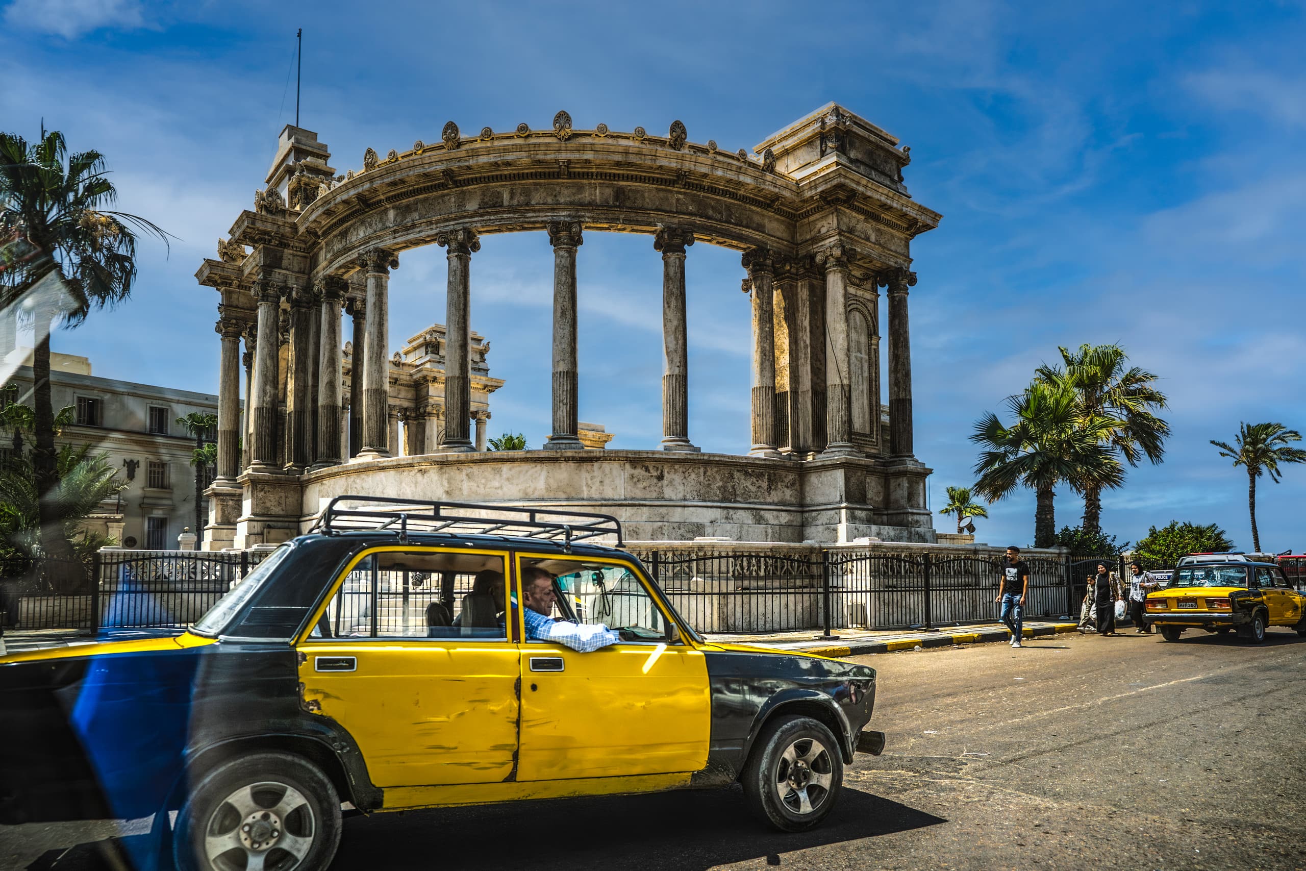 Street level view featuring a yellow and black taxi cab driving across a paved road, passing an ornate semicircular stone monument with towering classical columns and palm trees under a bright blue sky.