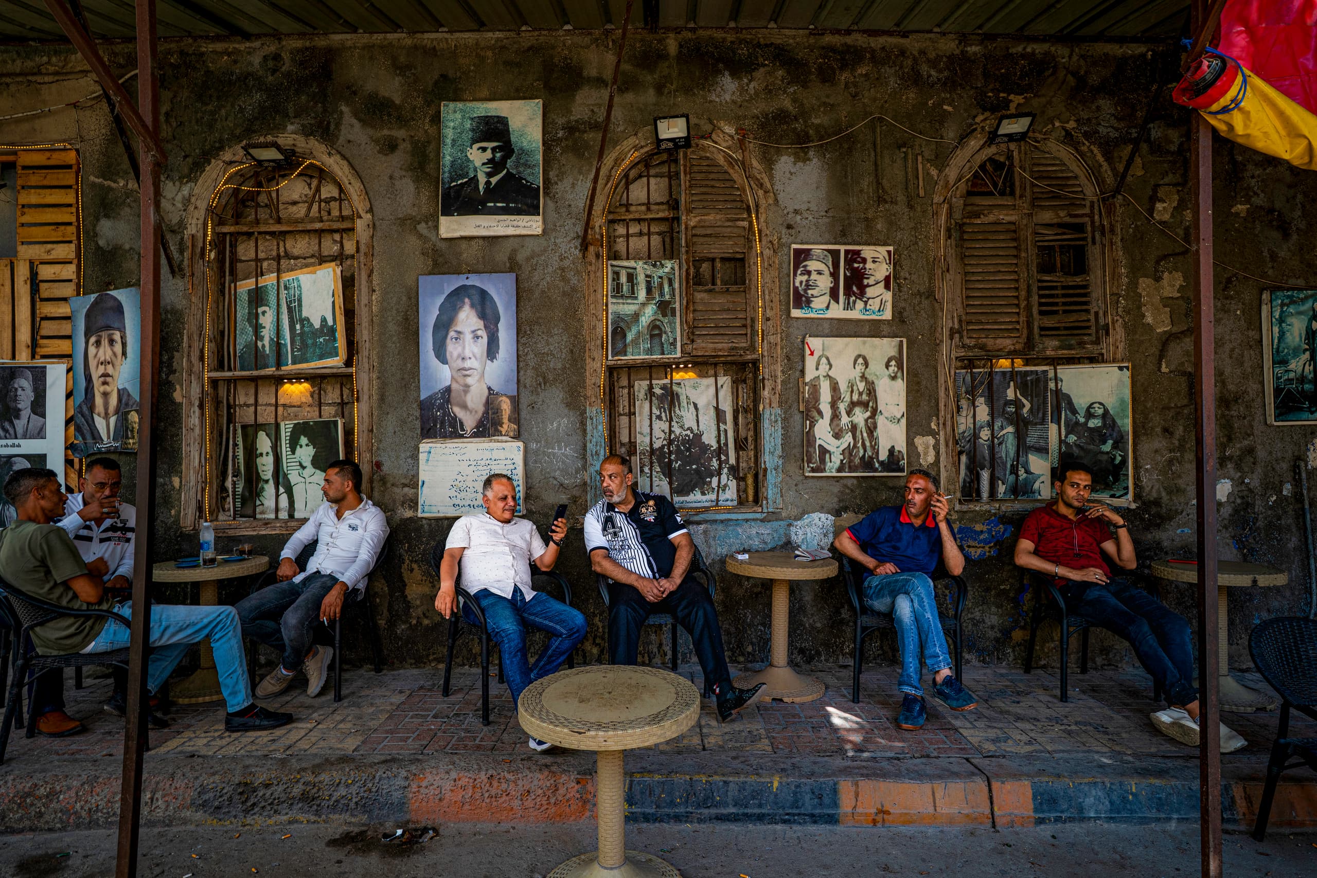 A wide shot of several men sitting along a textured wall decorated with old portraits and arched windows, each seated at a small round table under an overhang.