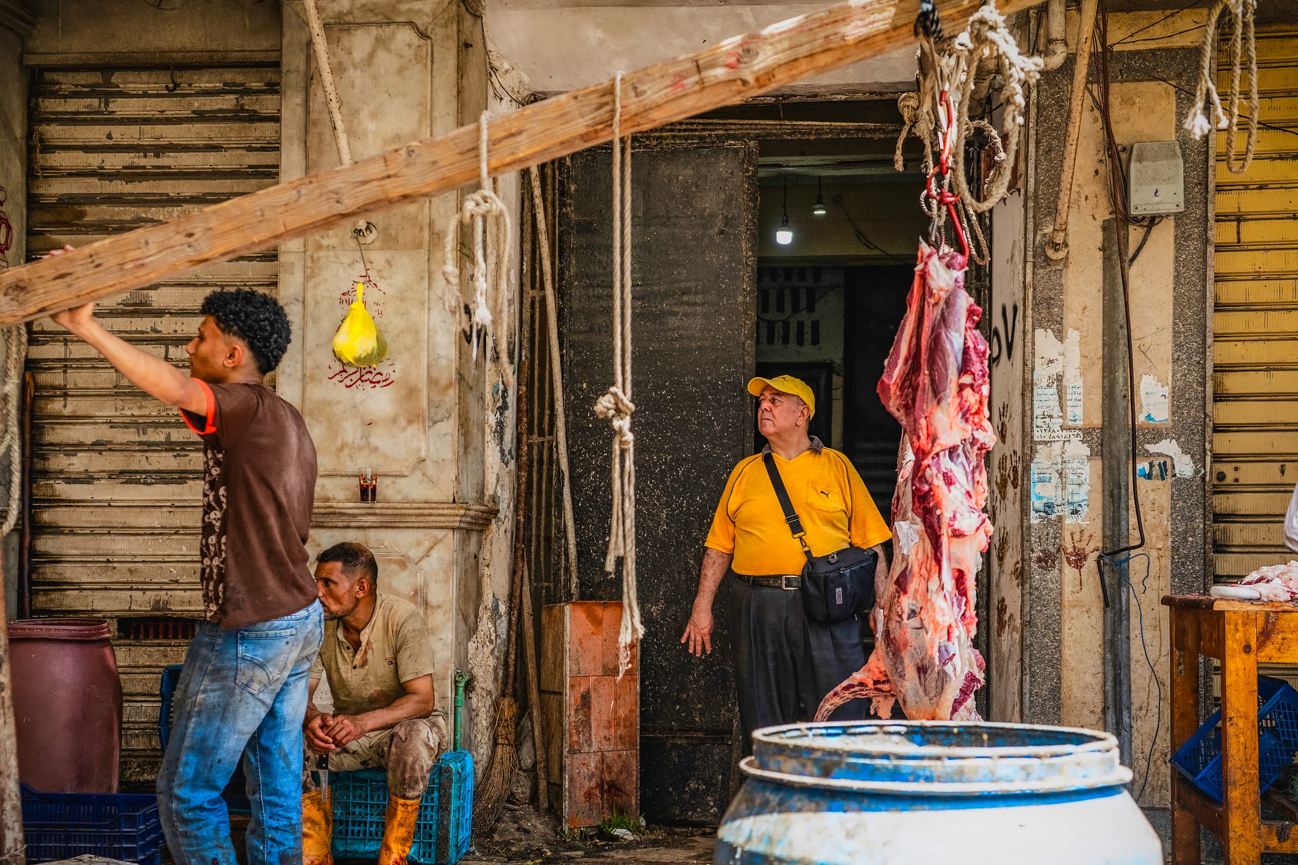 Street-level view of a butcher shop entrance with a man in a yellow cap and shirt standing inside, a seated worker in the foreground, another man lifting a wooden beam on the left, and a large piece of hanging meat in the right foreground.