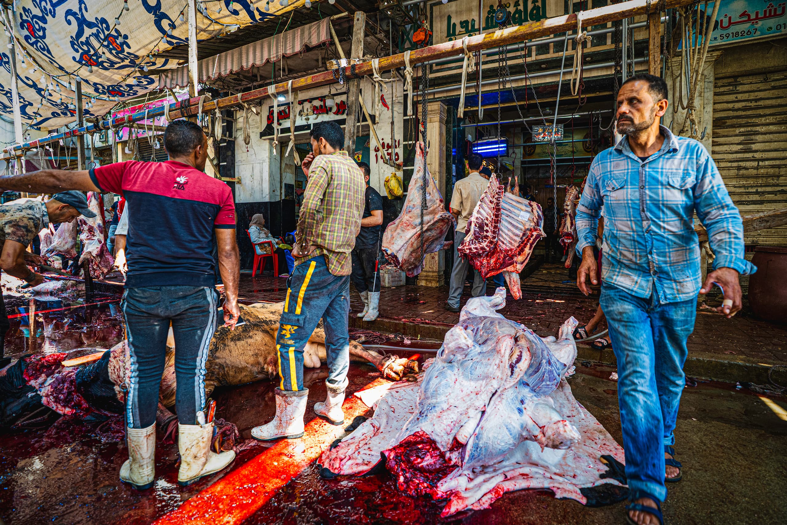 Wide-angle street scene of men working in a busy marketplace next to hanging and skinned animal carcasses on a wet floor, with a man in a blue plaid shirt walking in the right foreground.