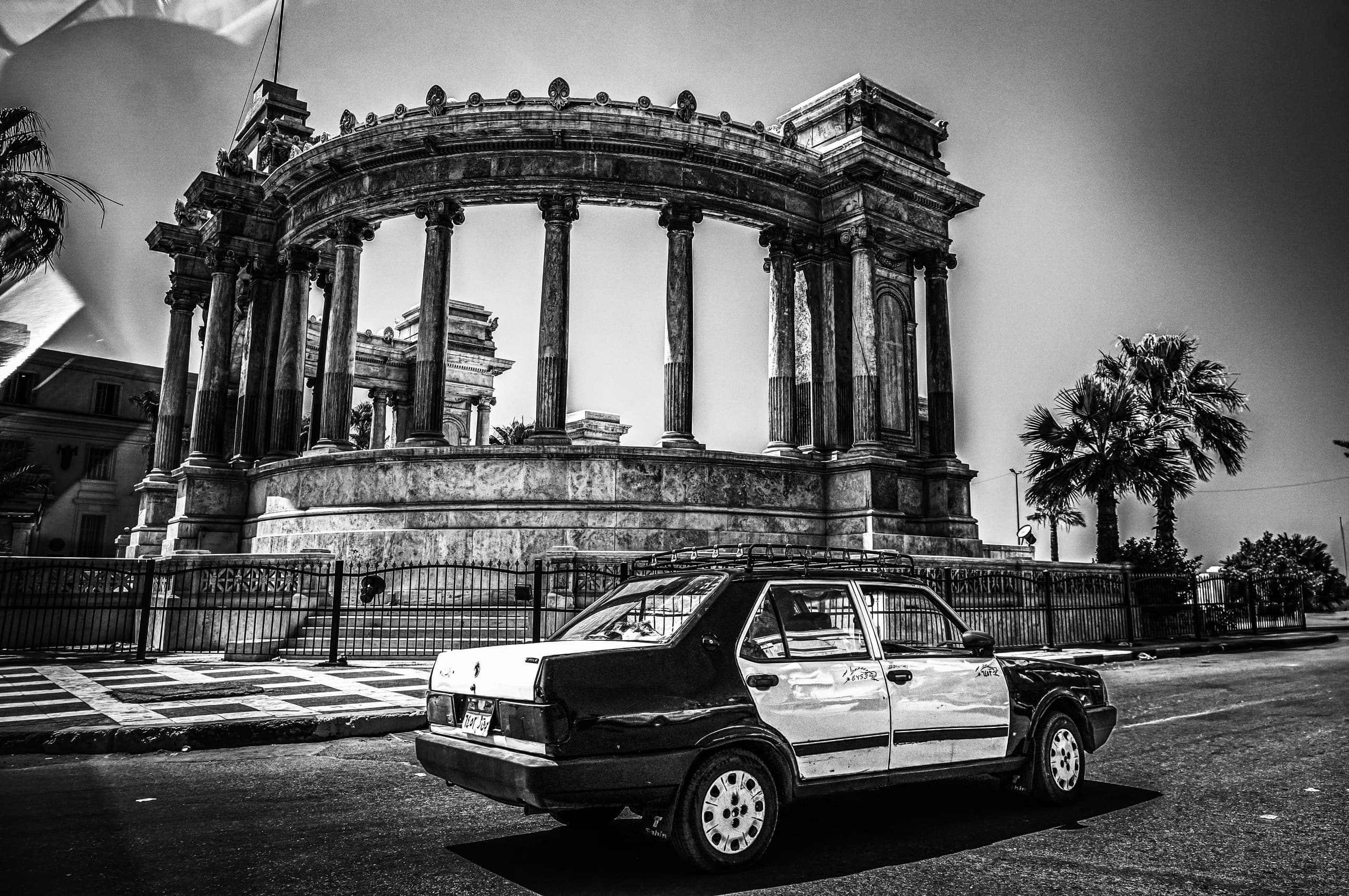 A black-and-white street-level photograph showing the rear profile of a two-tone taxicab parked on asphalt in front of a grand circular classical monument featuring tall stone columns and ornate cornices.