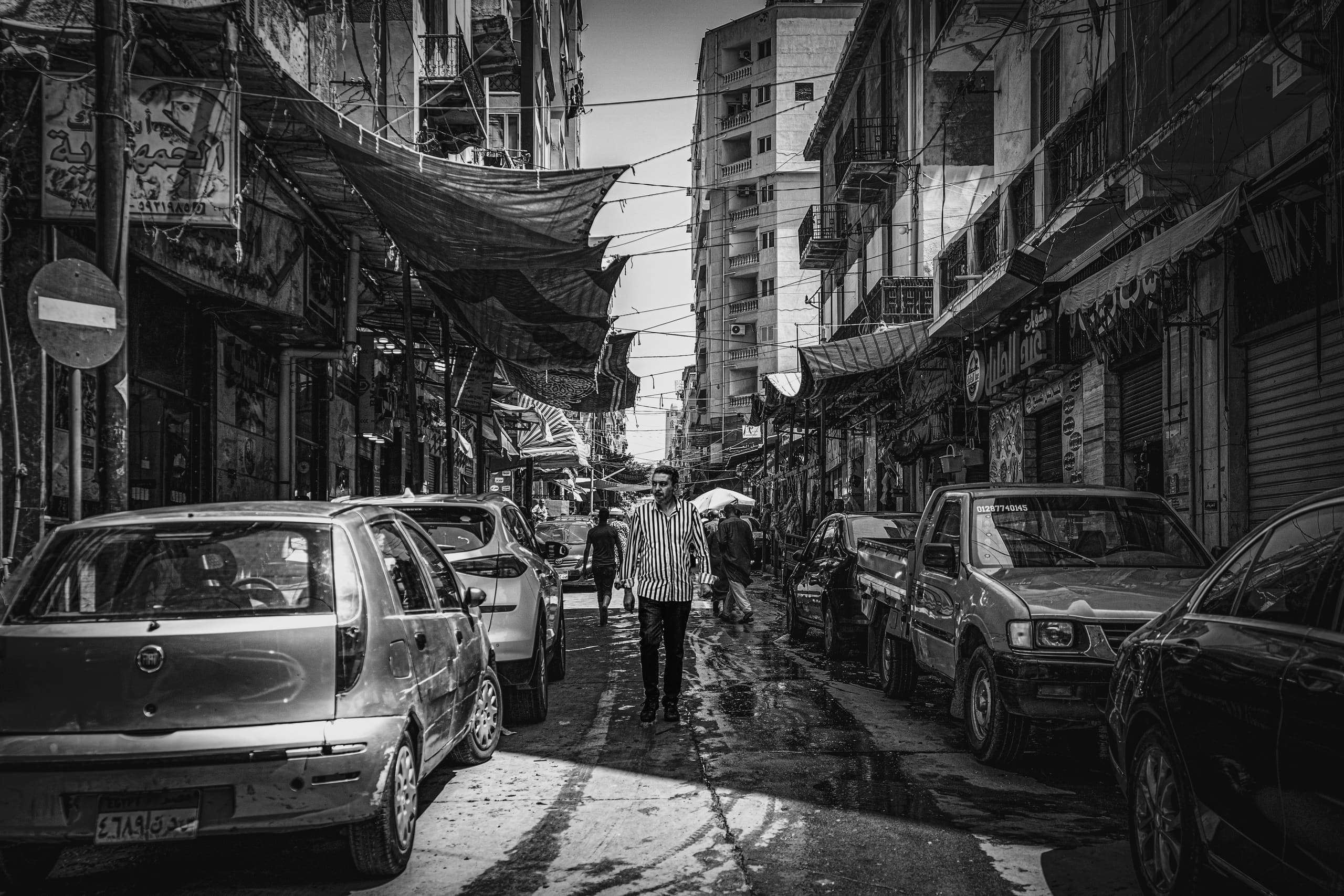 Black and white photograph of a man in a striped shirt walking down a narrow street lined with parked cars, overhead awnings, and tall buildings.