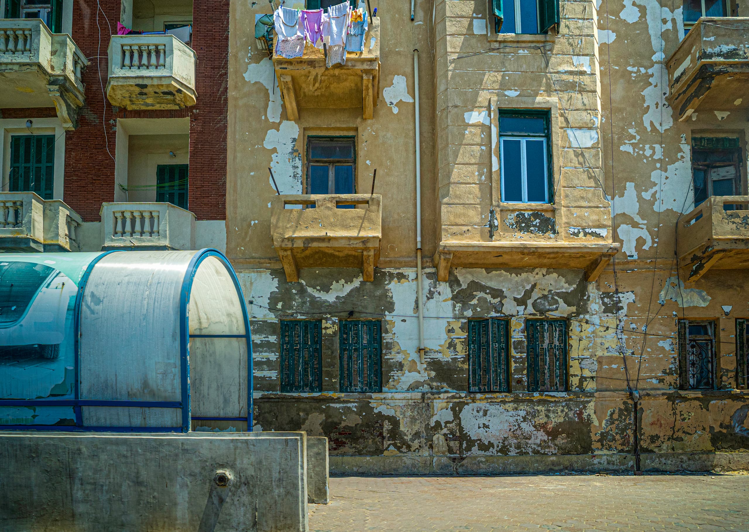 Exterior architectural view of a weathered multi-story building facade with peeling plaster, wooden shutters, small balconies, and hanging laundry, alongside a curved blue structure in the lower left foreground.