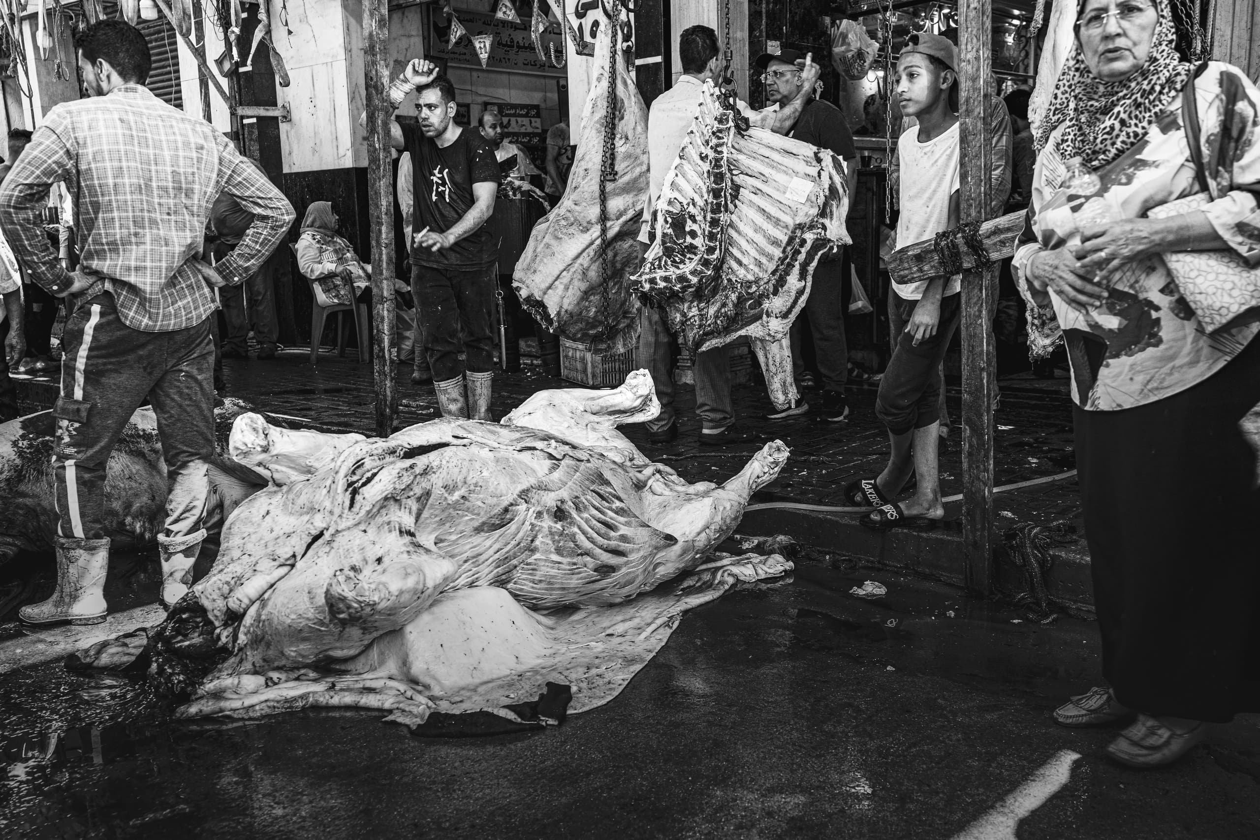 Monochrome street-level photograph of a busy meat market with a large butchered carcass on the wet floor in the foreground, hanging carcasses above, and several individuals standing around.