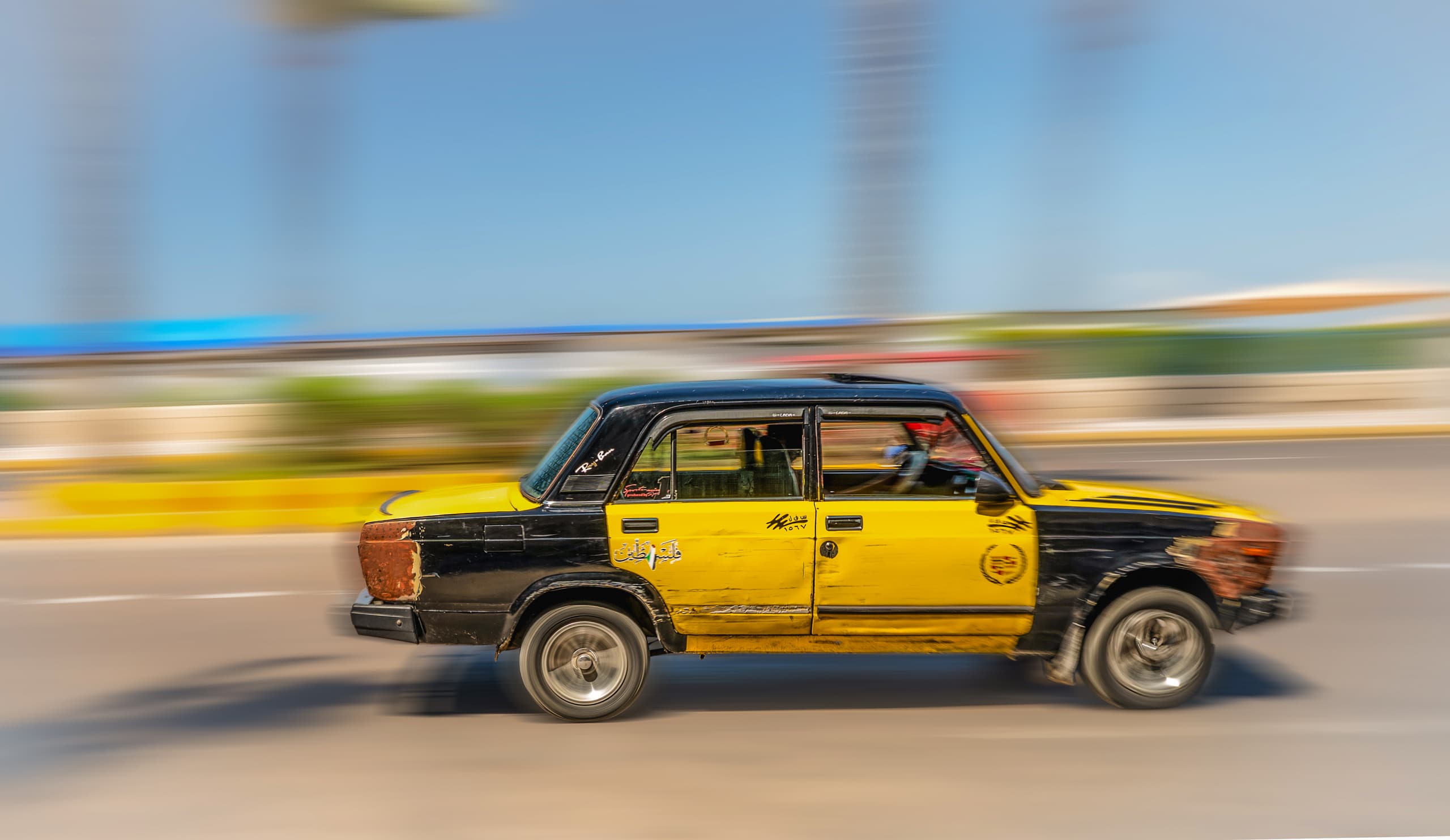 A panning shot of a weathered yellow and black car moving rapidly across an asphalt road against a blurred background under a clear sky.