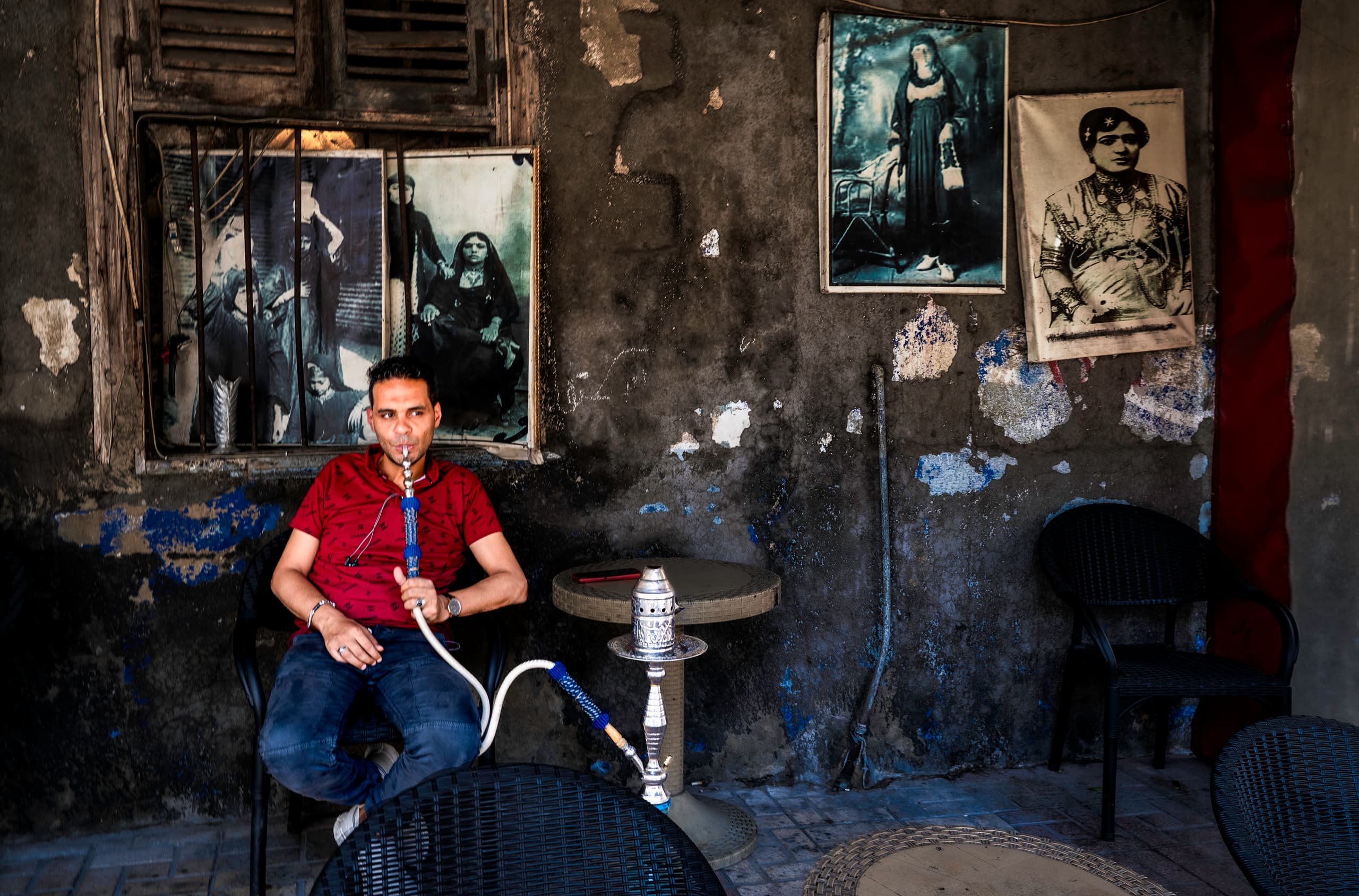 A seated man wearing a red shirt smokes a shisha pipe next to a small table, surrounded by peeling walls decorated with vintage portraits.