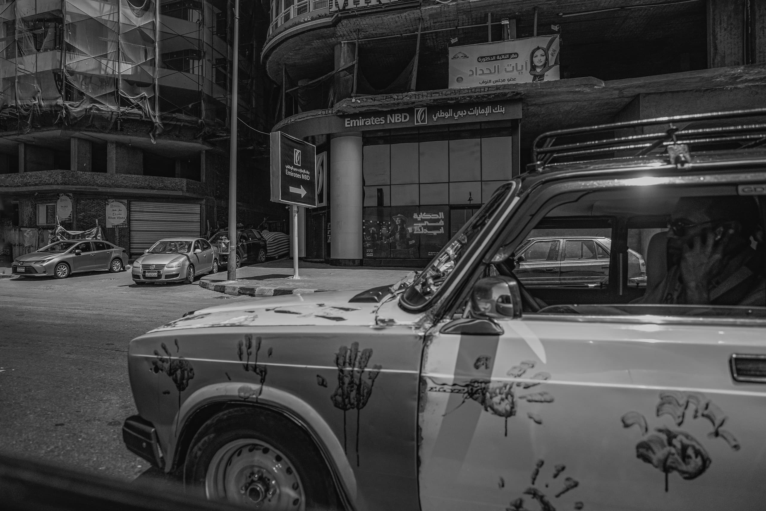 Monochrome street view focusing on the textured side and hood of a car covered in handprint marks in the foreground, with a driver on a phone inside, parked cars and commercial buildings across the street in the background.