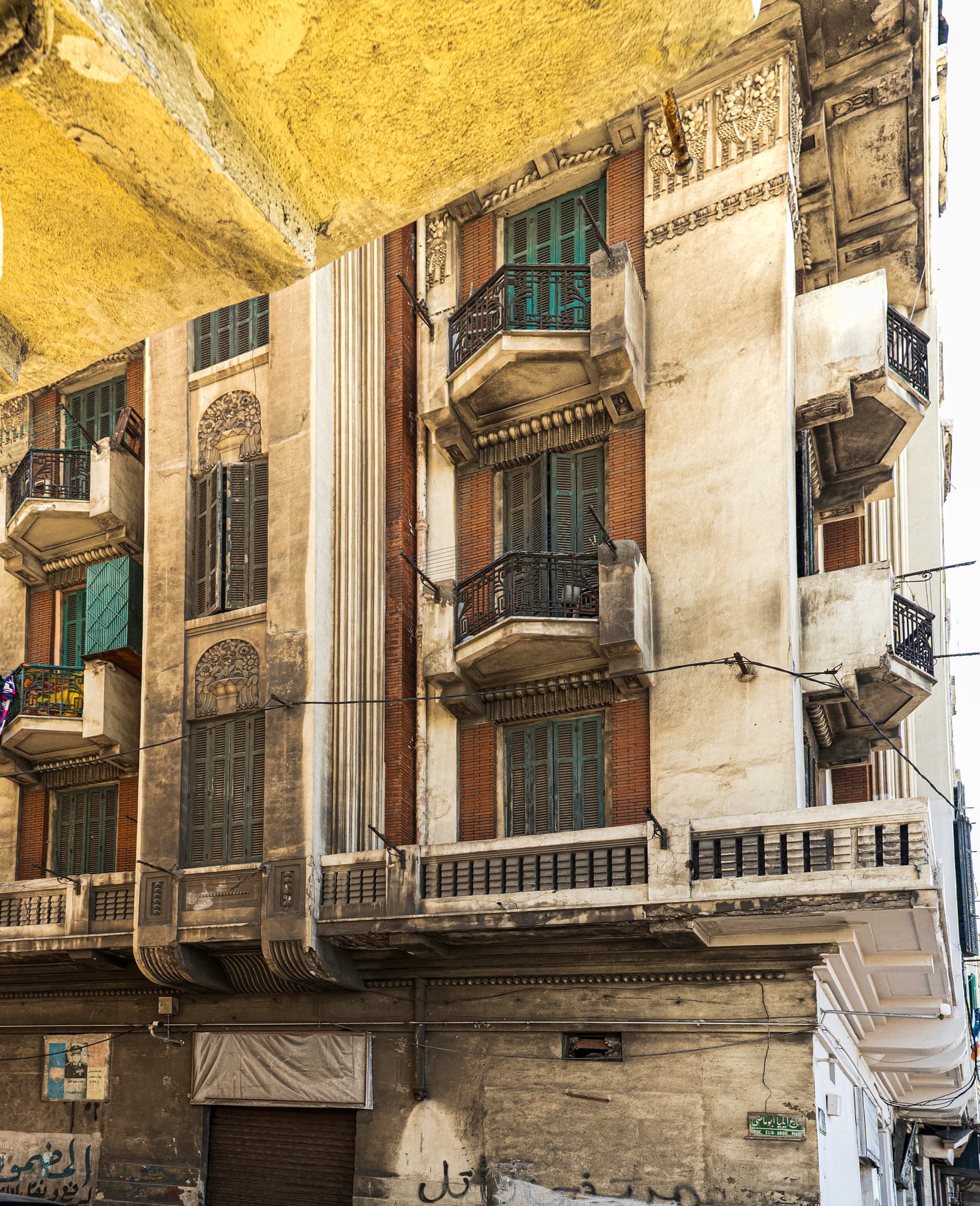 A low-angle view of a weathered urban building facade showing stacked balconies with dark metal railings, teal louvered window shutters, exposed red brickwork, and aged concrete surfaces under daylight.