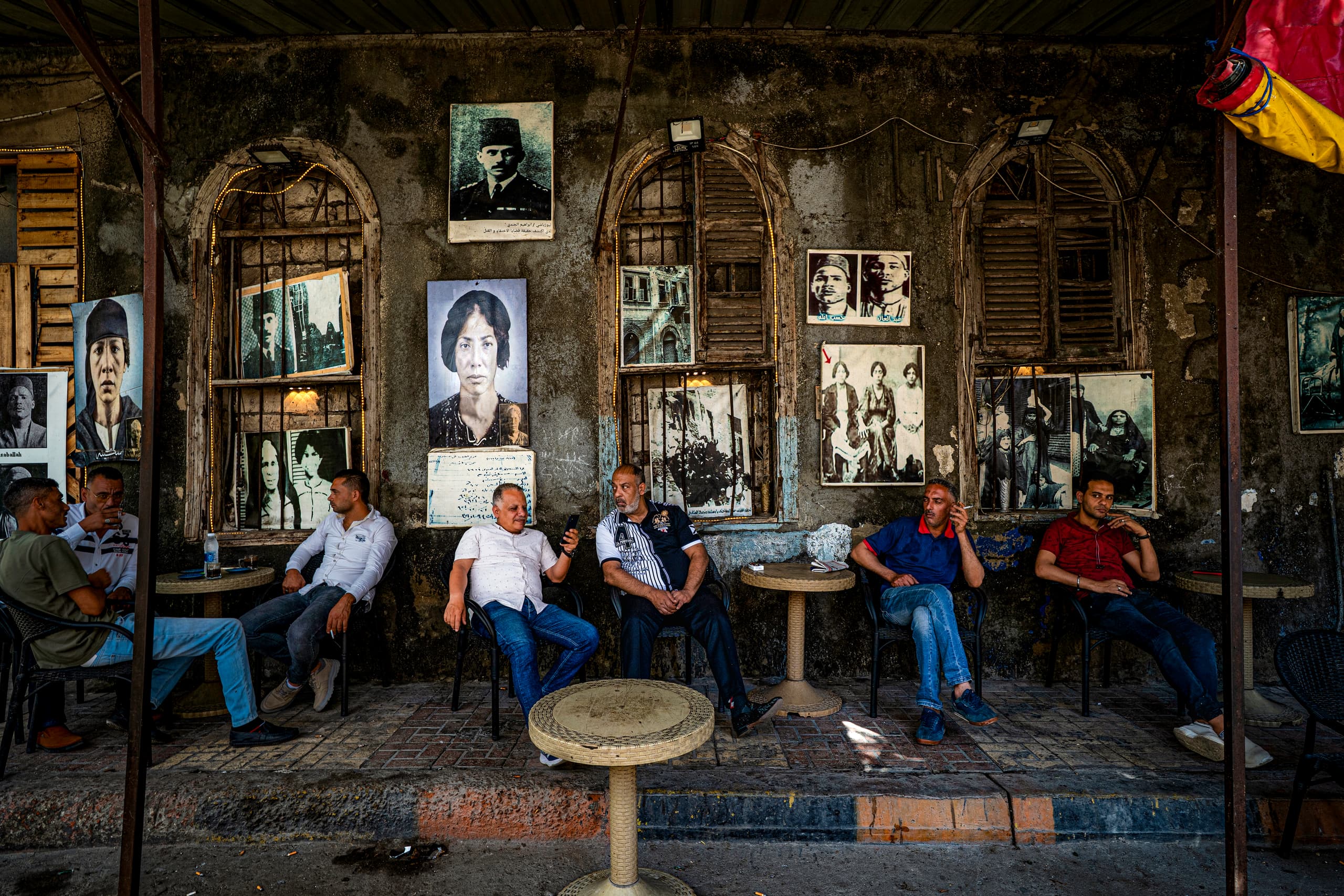 Horizontal color photograph showing six men sitting along a textured stone wall lined with old portraits and arched shuttered windows, separated by small plastic tables on a paved sidewalk.