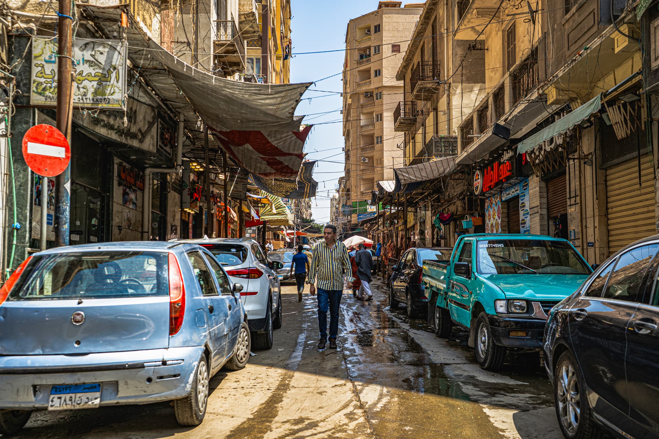 A man in a striped yellow shirt walks down the middle of a tight street flanked by parked cars on both sides and old buildings overhead, captured in bright daylight.