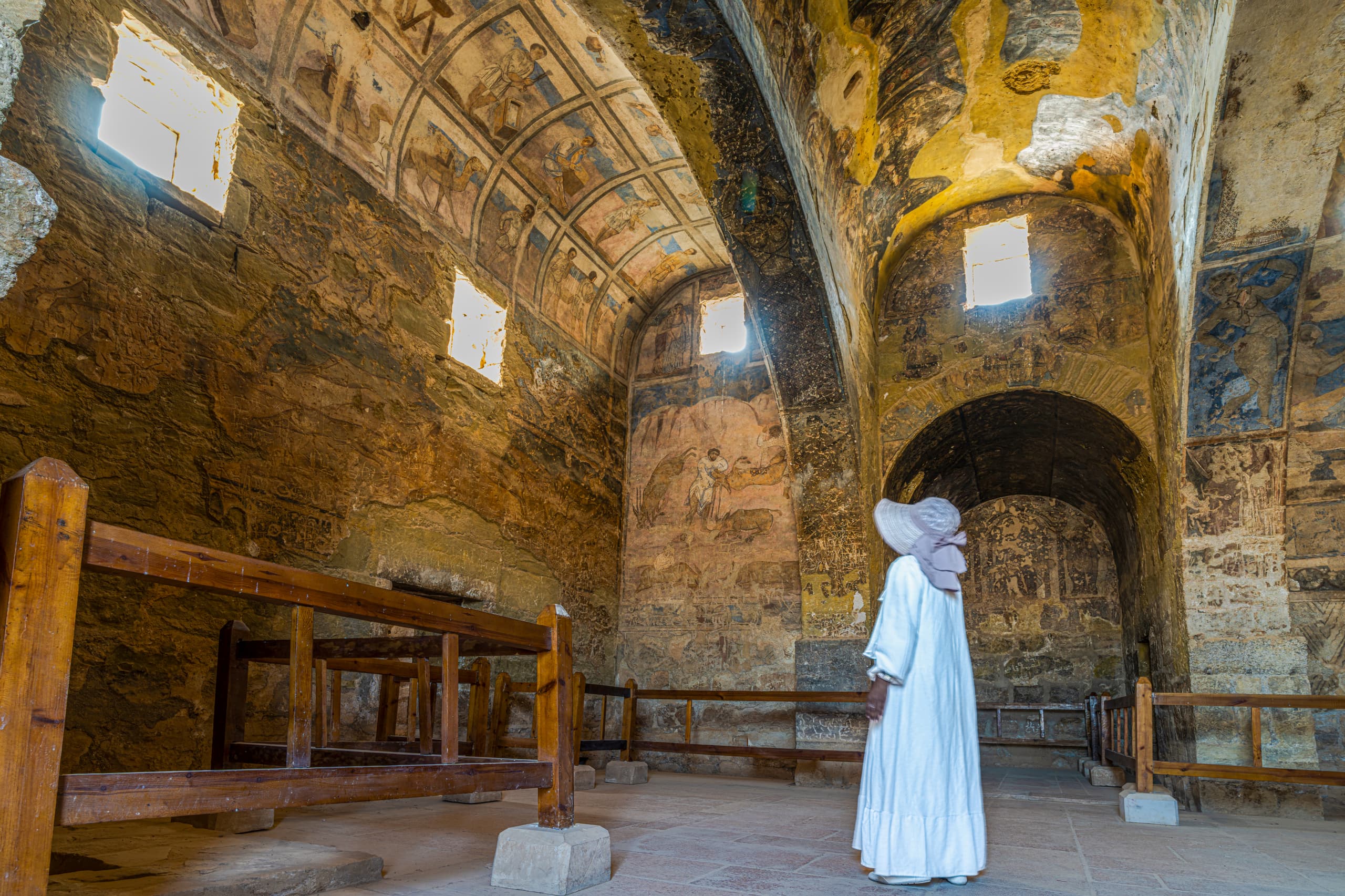 A person wearing a white dress and hat stands inside a vaulted stone chamber, looking upward at weathered frescoes on the ceiling and walls, framed by low wooden barriers in the foreground.