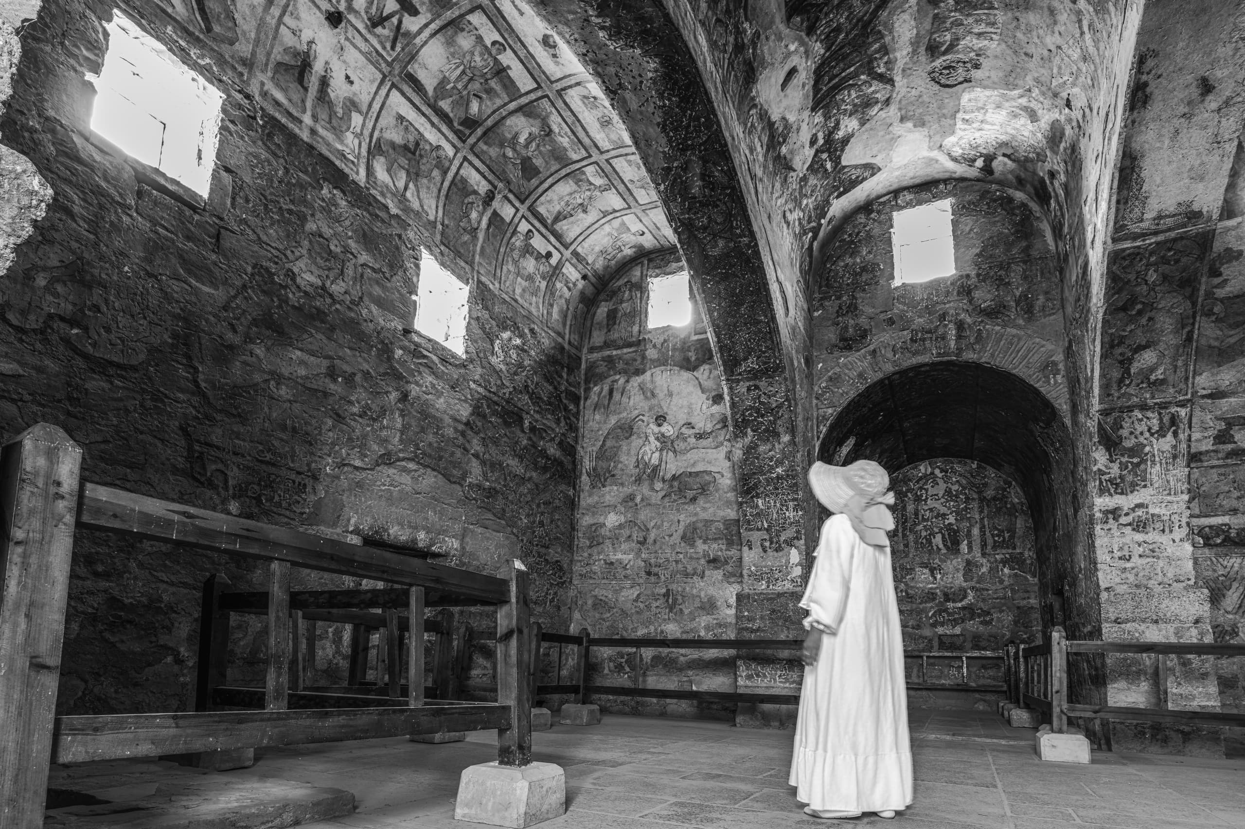 A full-length monochrome photograph of a figure in a white dress and wide-brimmed hat standing inside a vaulted stone hall with ancient frescoes, looking upward toward murals and high rectangular windows.