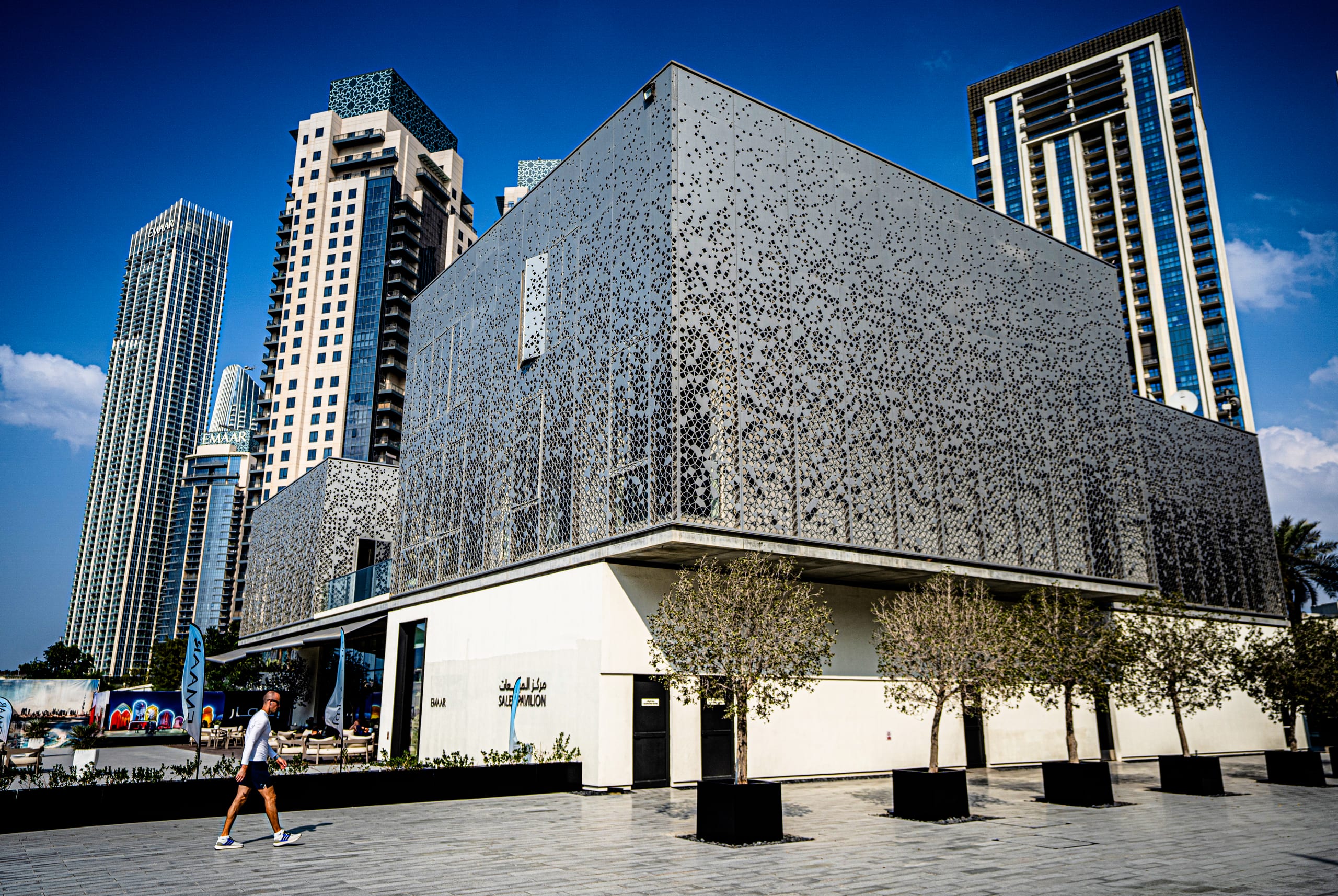A man in a white long-sleeve shirt and dark shorts walks left along a paved plaza, passing a modern multi-story pavilion with a decorative perforated facade and four potted trees, with multiple skyscrapers rising in the background under a blue sky.