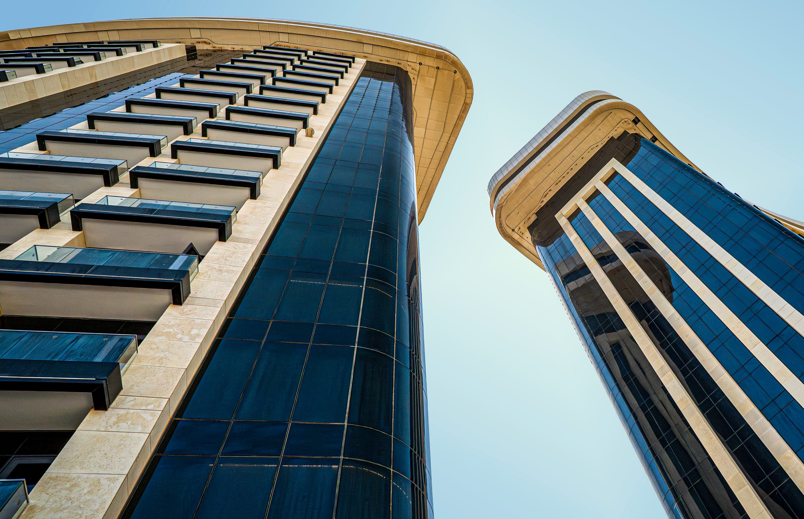 Low-angle architectural shot looking straight up at two modern high-rise towers with curved facades, dark reflective glass, and light-colored stone trim against a clear blue sky.