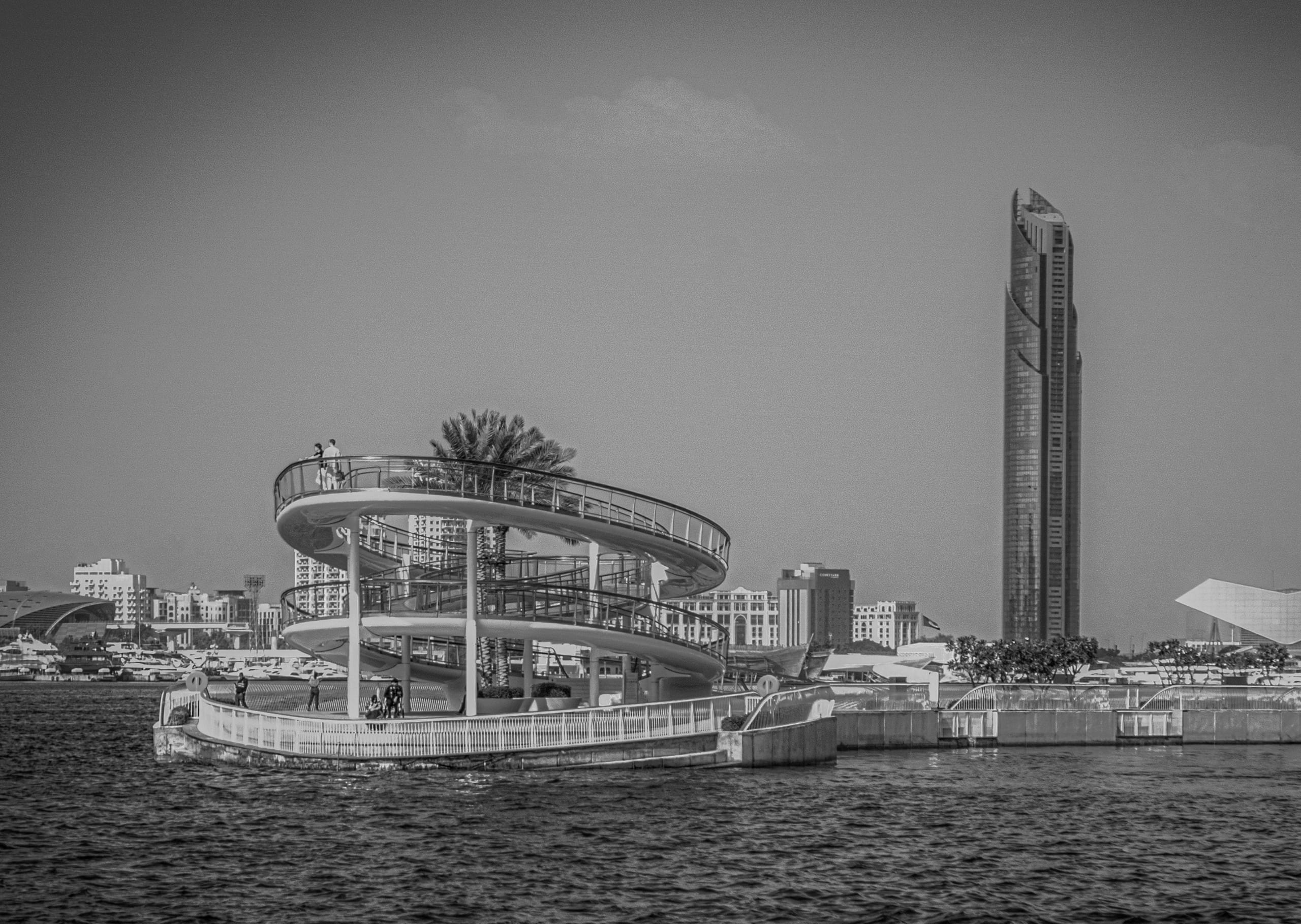 A black-and-white architectural photograph showing a multi-tiered spiral observation structure sitting in the water, with a tall, slender skyscraper rising in the background and smaller figures visible on the ramps.