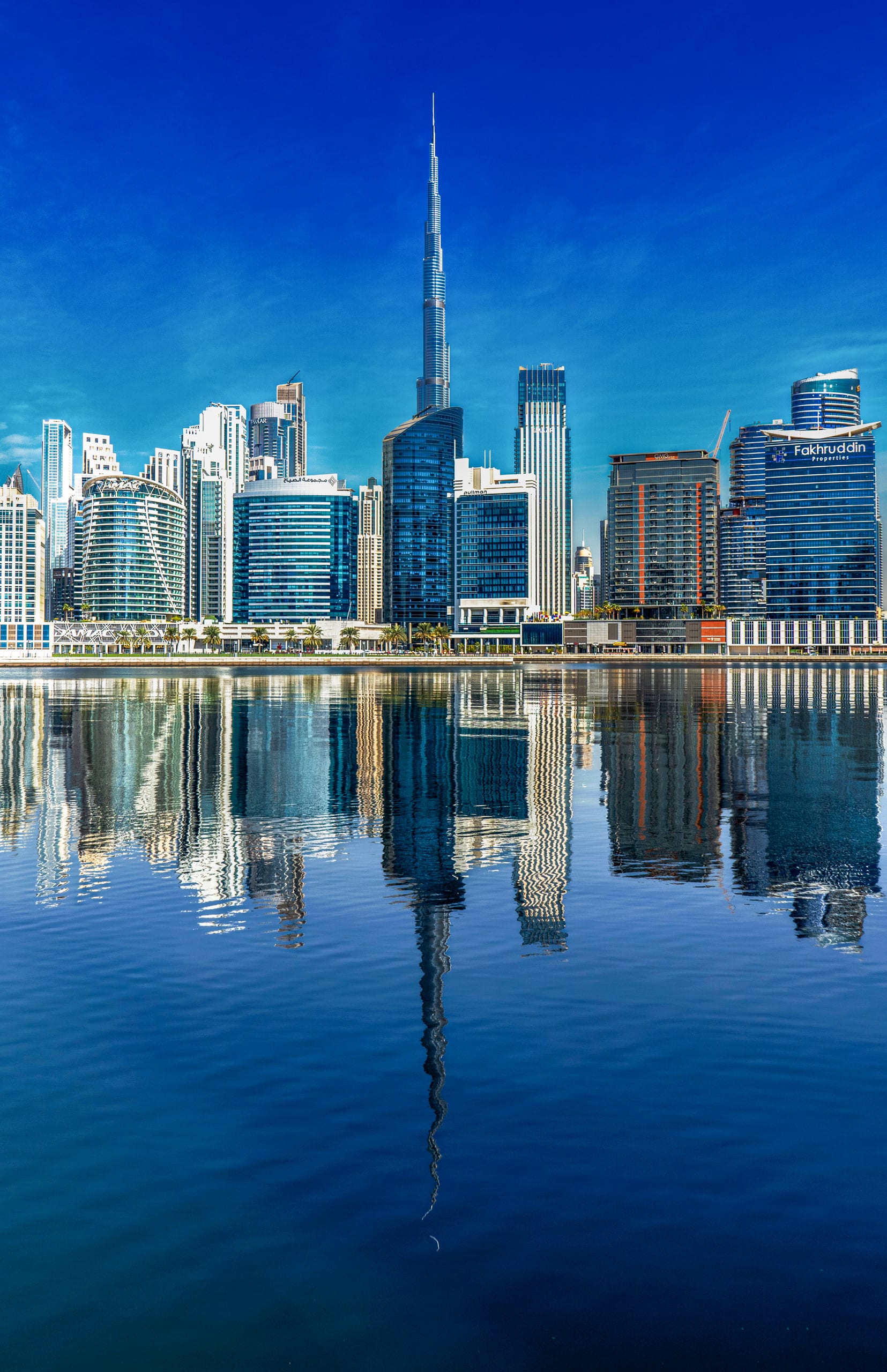 A vertical exterior view of a tall slender skyscraper rising above a dense cluster of modern high-rise buildings, with their complete reflections mirrored on the surface of calm water in the foreground under a clear blue sky.
