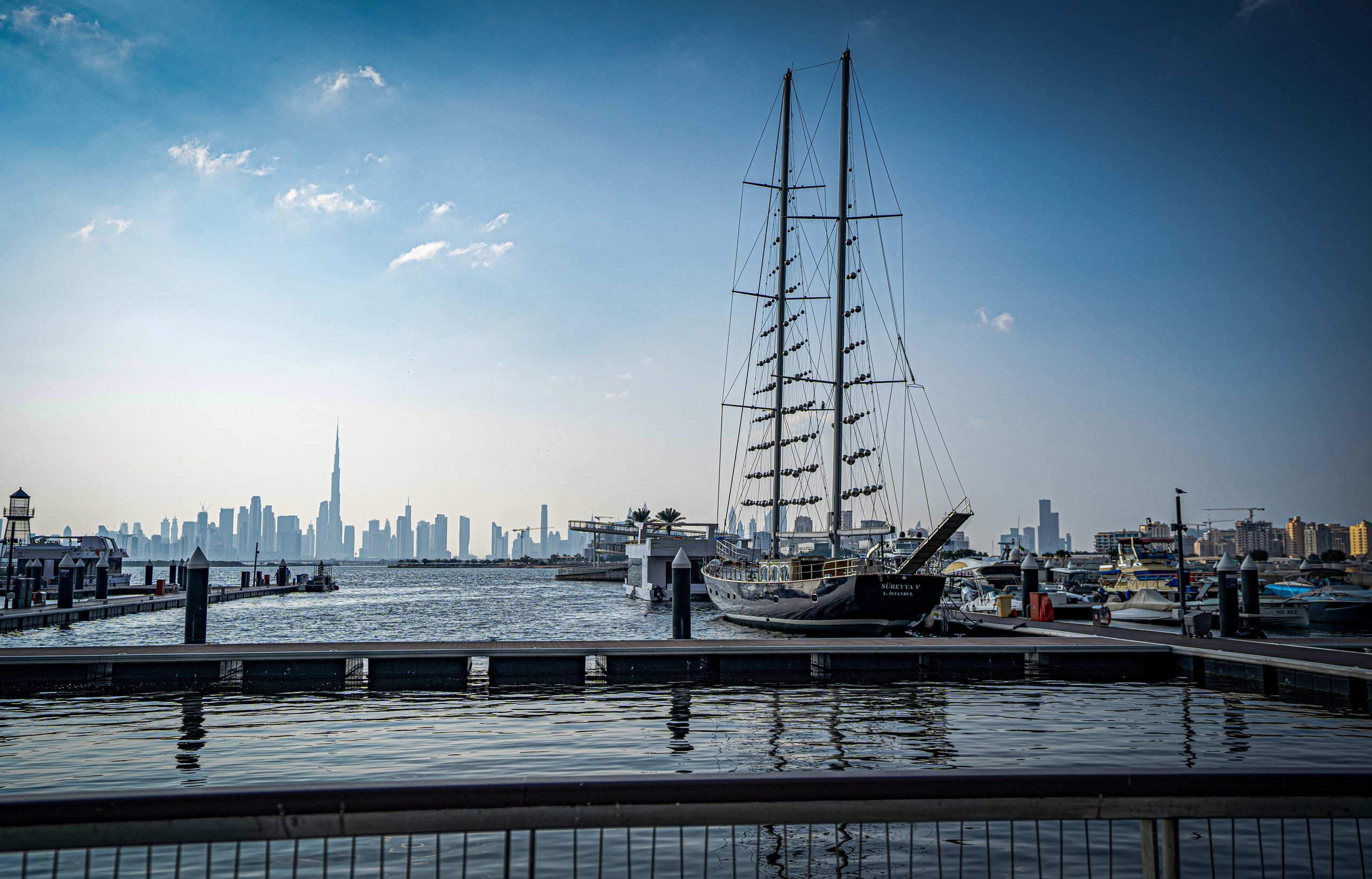 A large two-masted traditional sailing ship moored at a wooden and concrete marina pier, with a city skyline visible across the water in the hazy background under a bright sky.