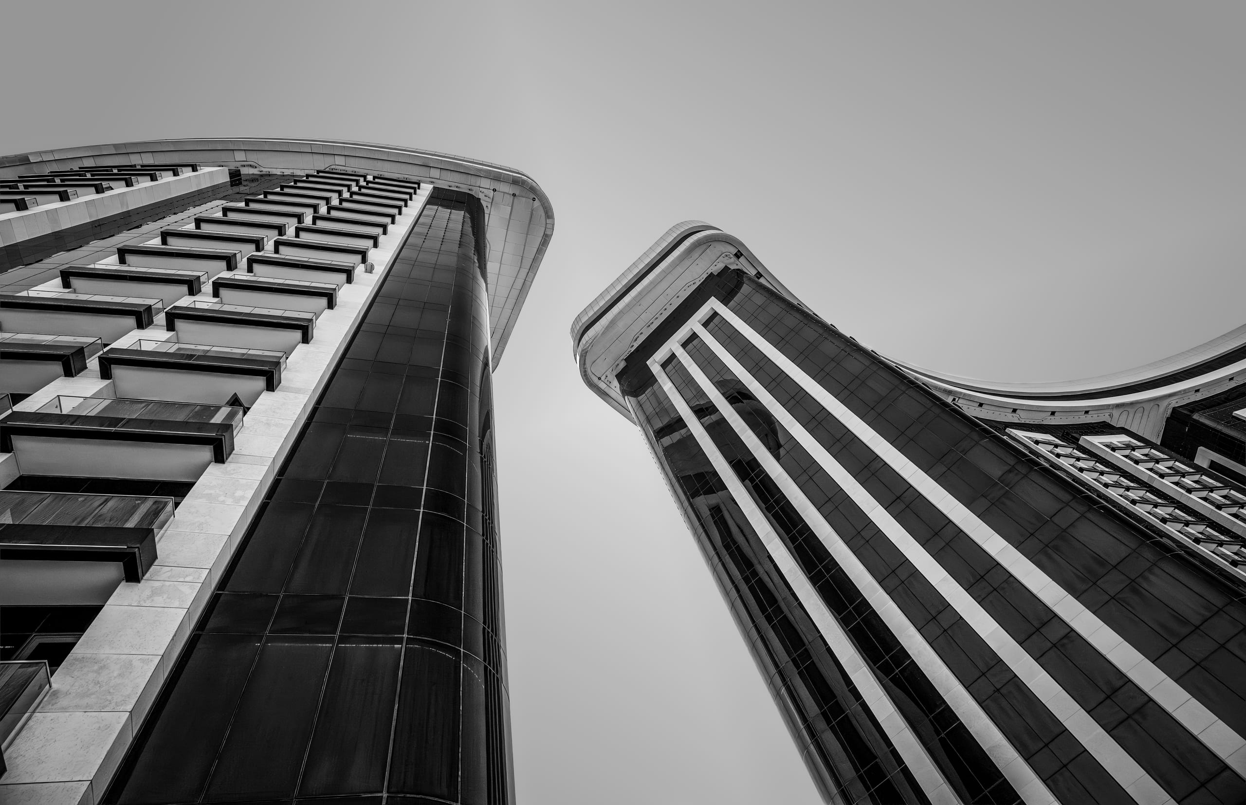 A low-angle black-and-white architectural photograph looking straight up at two modern curved skyscrapers with dark glass windows and contrasting white structural ribs, separated by a slice of pale sky.