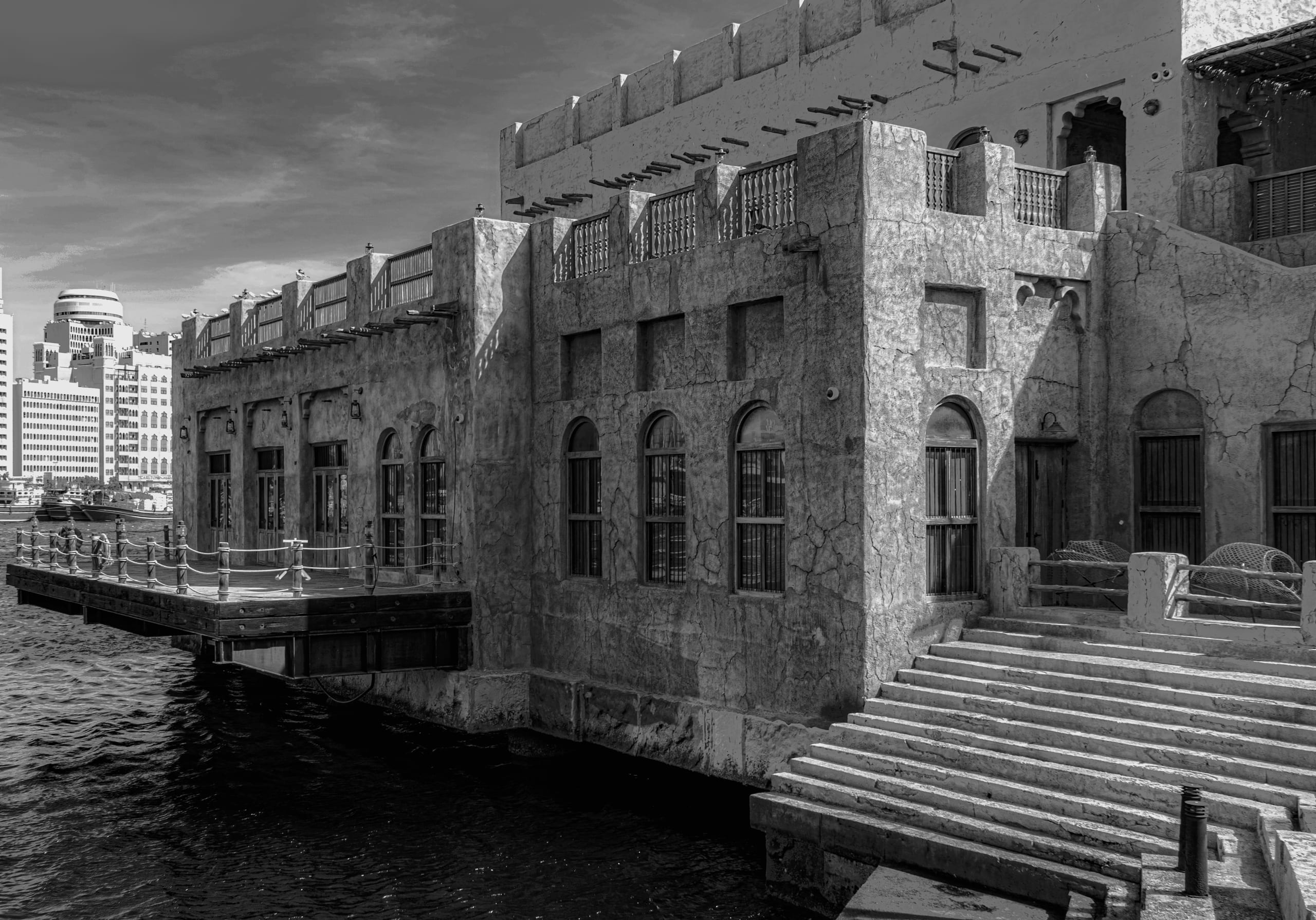 Monochrome architectural photograph showing a historic multi-level stone building with traditional arched windows and a wooden boardwalk over dark water, contrasting with a modern skyscraper in the distant background.