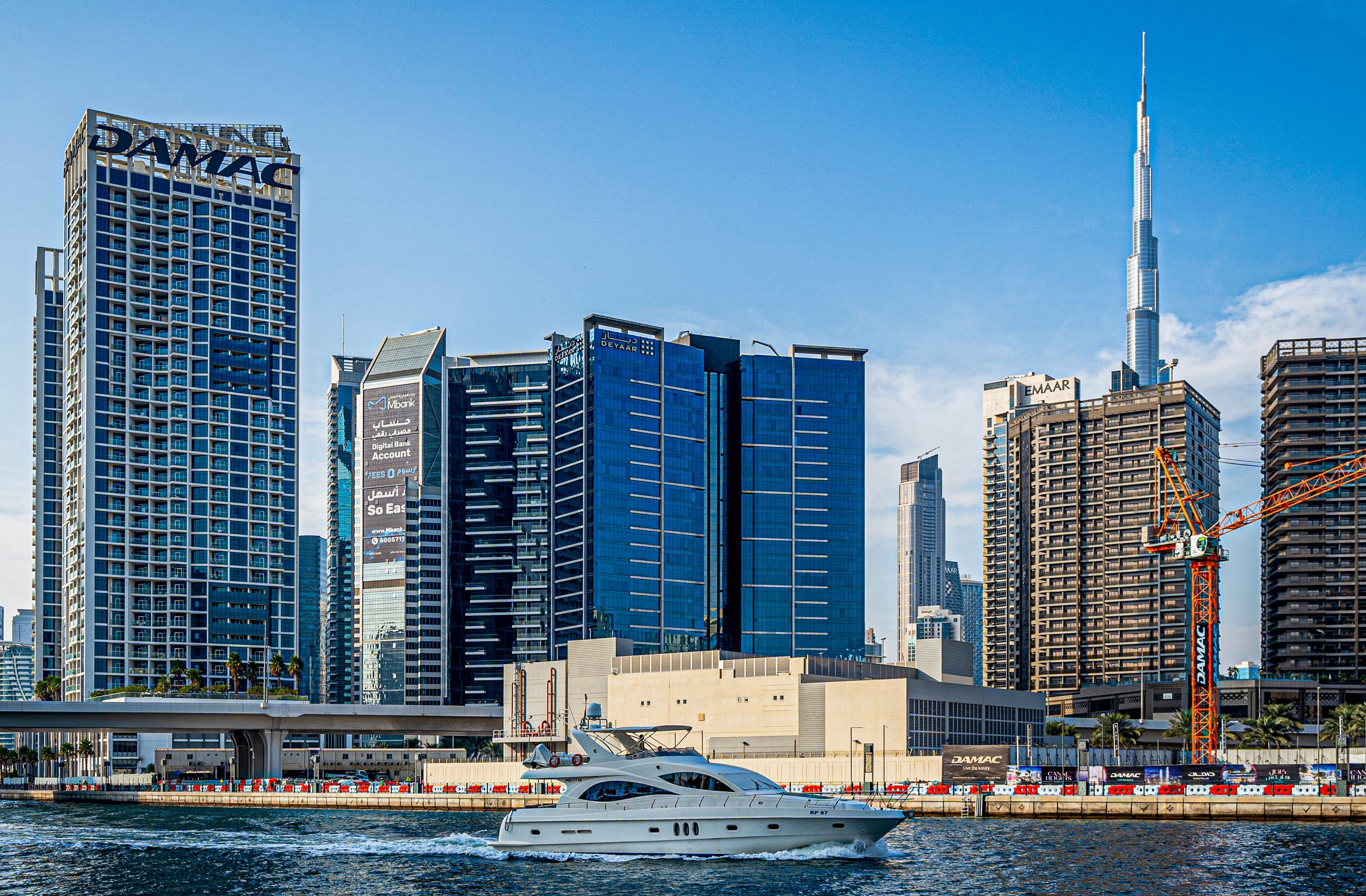 A white yacht moving across a blue canal water in front of a dense urban skyline featuring tall skyscrapers, modern buildings, and a bright clear sky.