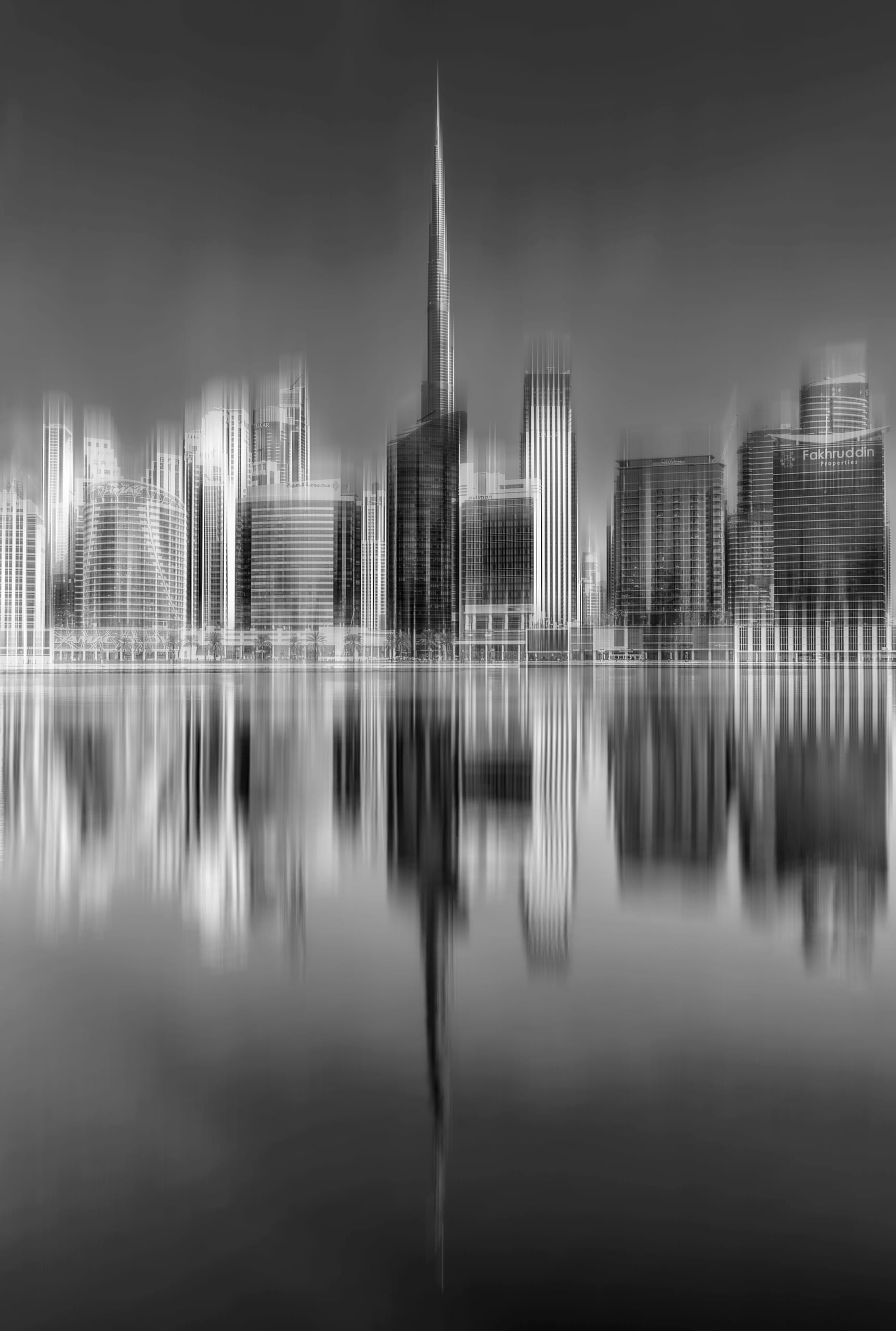 Black and white long exposure photograph of a high-rise city skyline across a calm waterfront, featuring vertical motion blur on the buildings and their sharp downward reflections in the water.