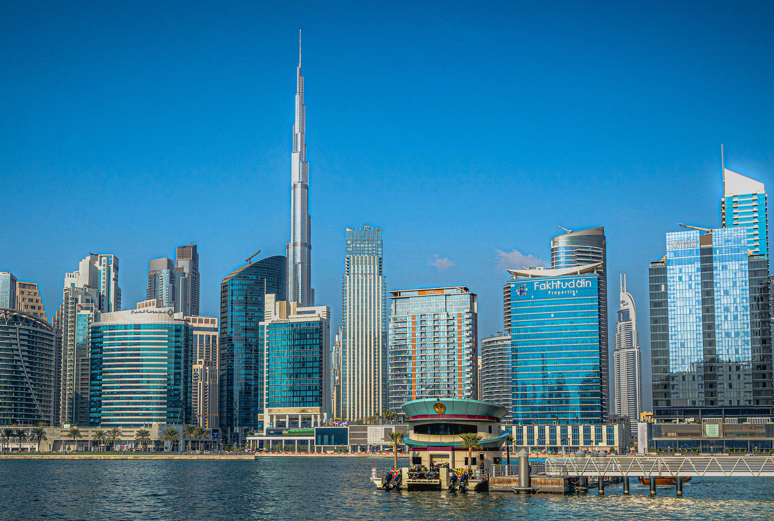 A wide daylight view of a modern city skyline dominated by a slender supertall skyscraper rising above a dense array of high-rise office and residential towers, with a calm waterway and a floating terminal building in the foreground.