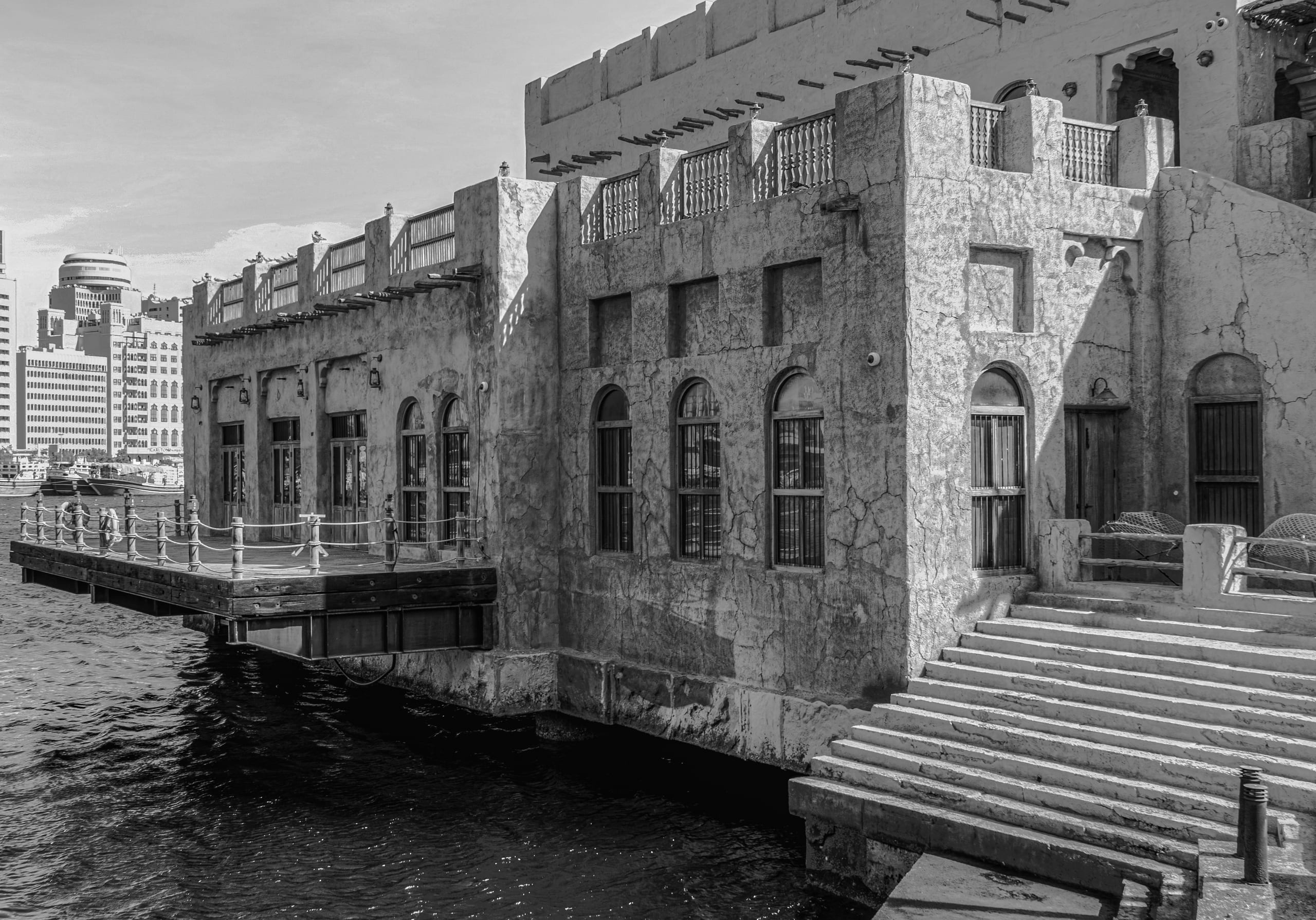 Monochrome architectural photograph showing low-rise textured concrete and stone buildings on a waterfront with wooden jetties and stairs, contrasted against modern skyscrapers in the distant background.