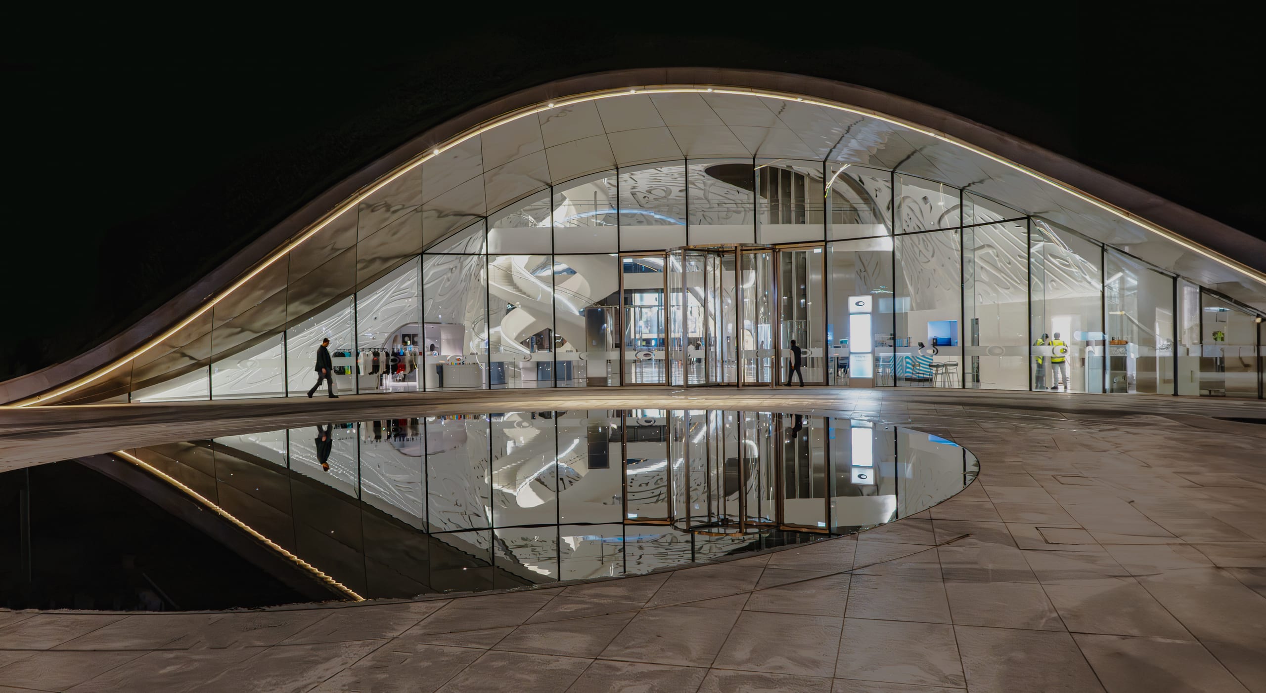 Night view of a low, wide modern building with a curved arch roof and glass facade, featuring a solitary person walking left and a puddle reflecting the illuminated entrance.