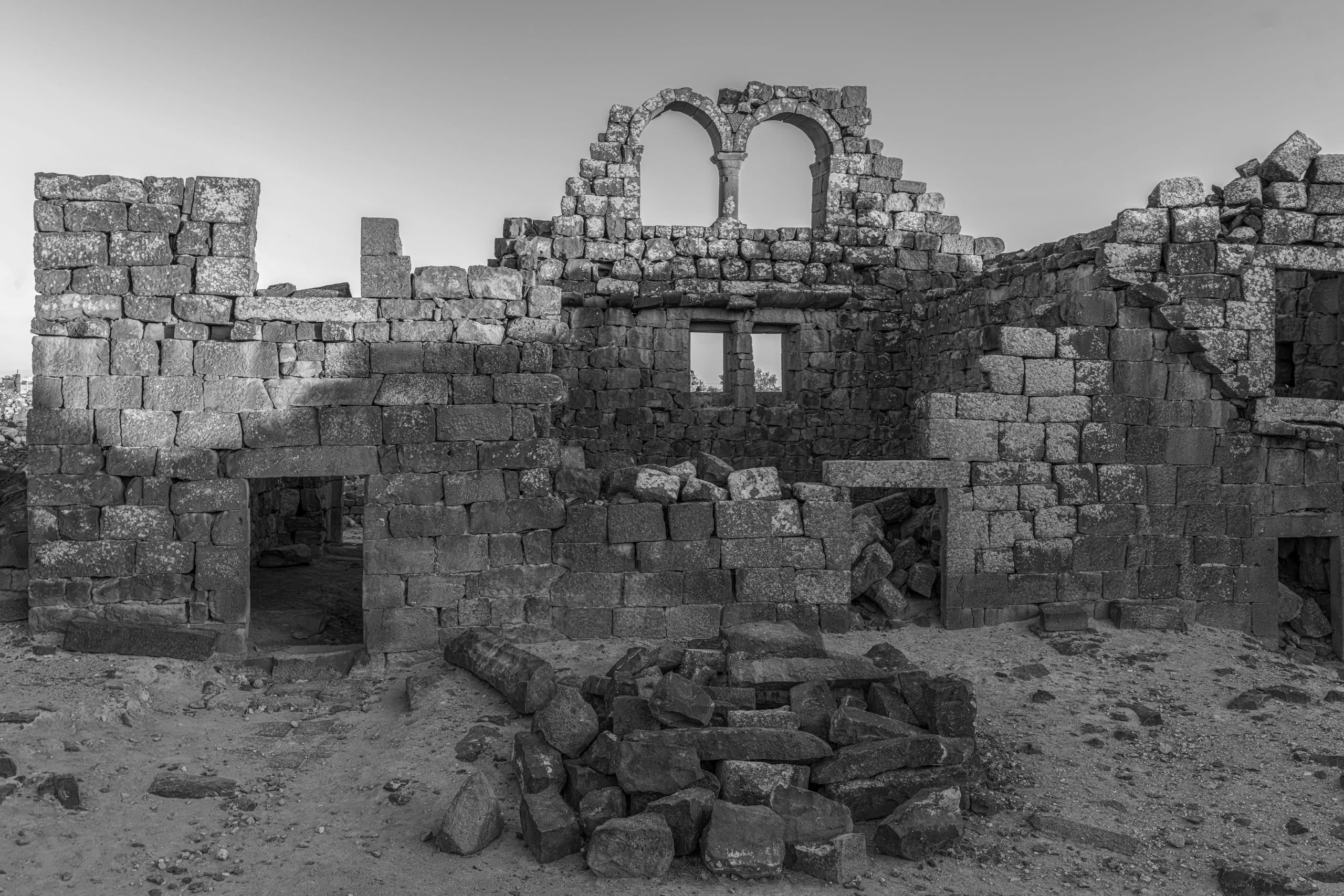 Black and white photograph of weathered stone ruins featuring an upper wall with two rounded arched window openings, rectangular doorways, and scattered rubble in the foreground under a plain sky.